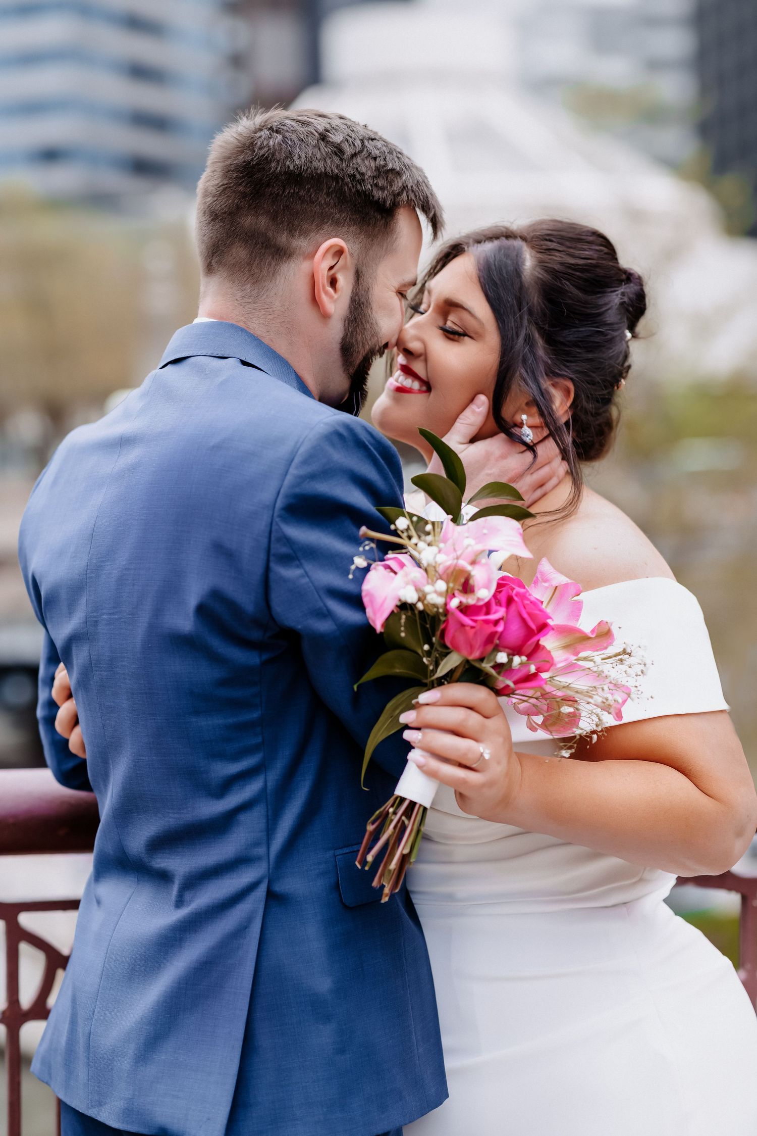 Groom tenderly kisses bride wearing white off-shoulder dress as she holds pink peony bouquet on a city bridge.