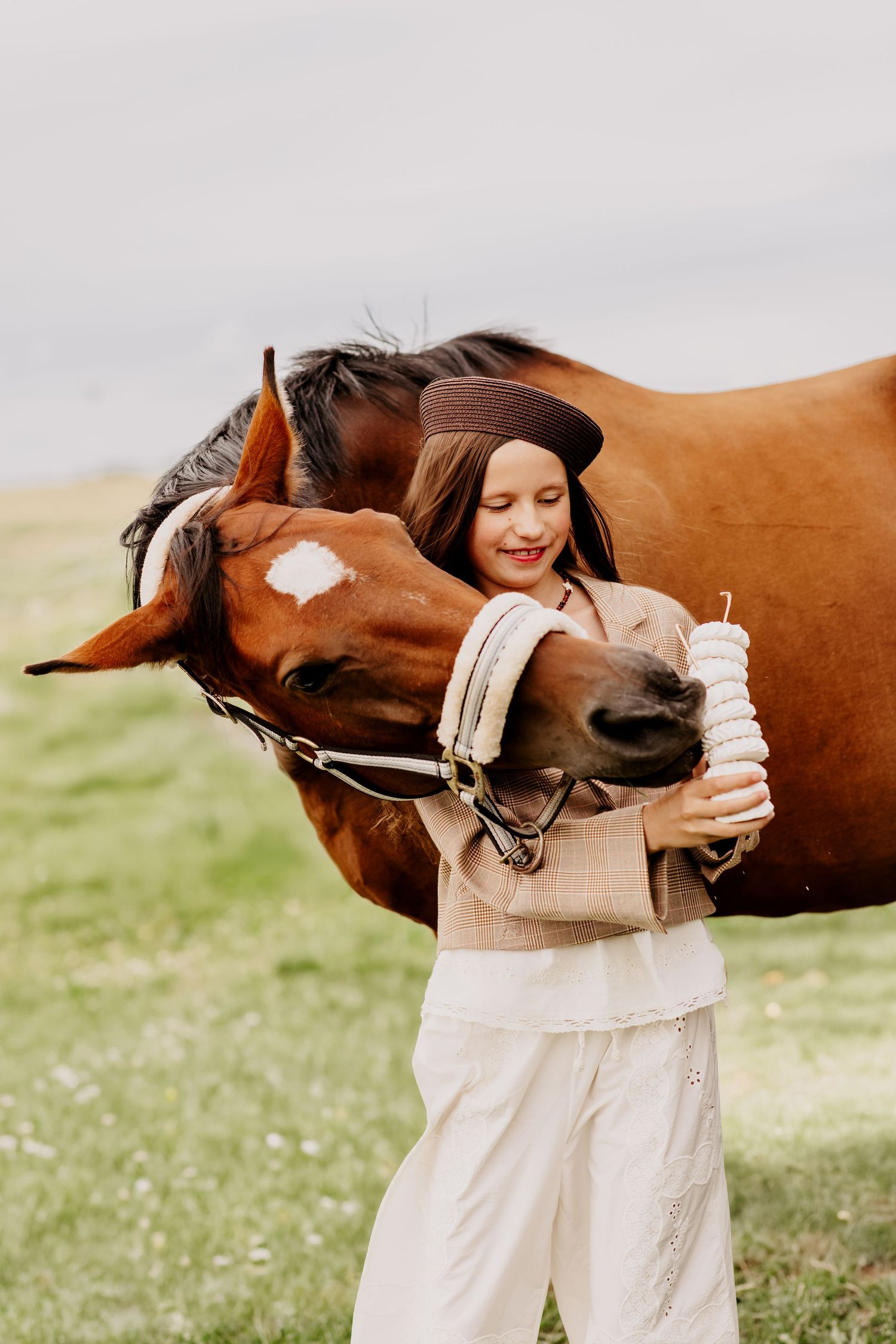 Laughing girl in brown beret hugging a chestnut horse while holding white stacked treats in a green meadow.