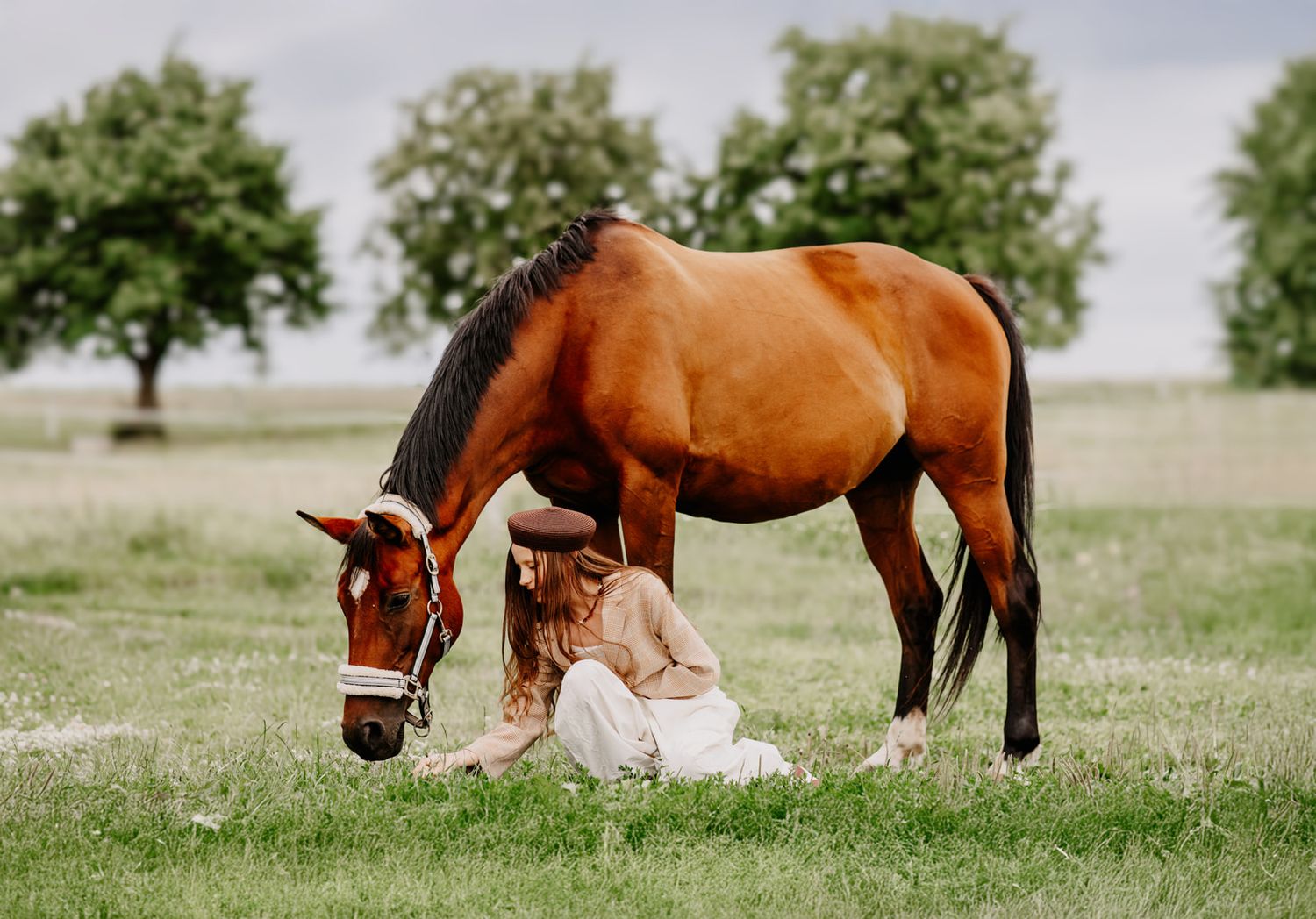 Woman in white dress sits beside a grazing bay horse in a lush green field.