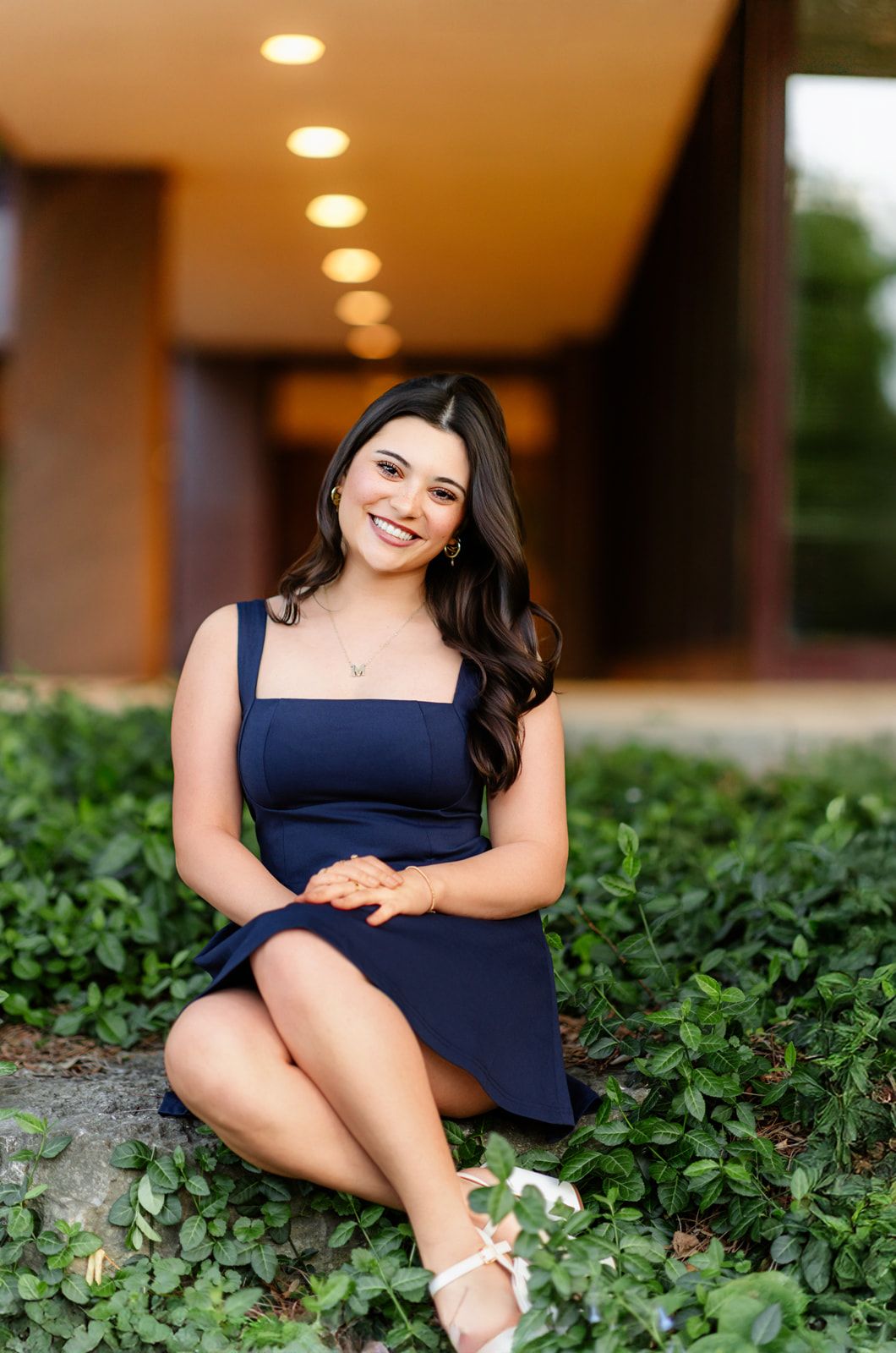 Smiling young woman in navy blue dress sitting on ground among green clover plants.