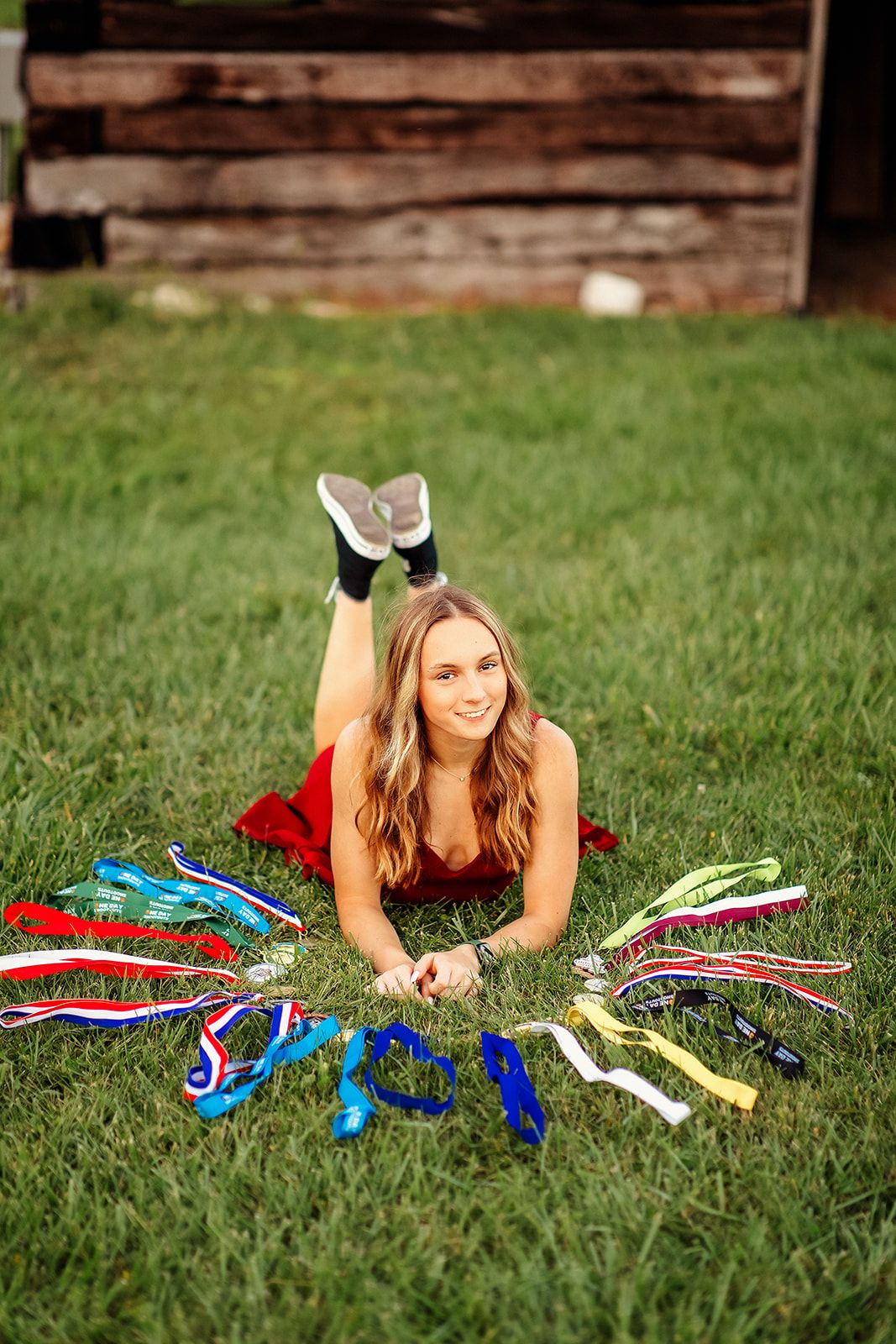 Teen girl in red dress lying on grass surrounded by colorful medals arranged in a fan shape.
