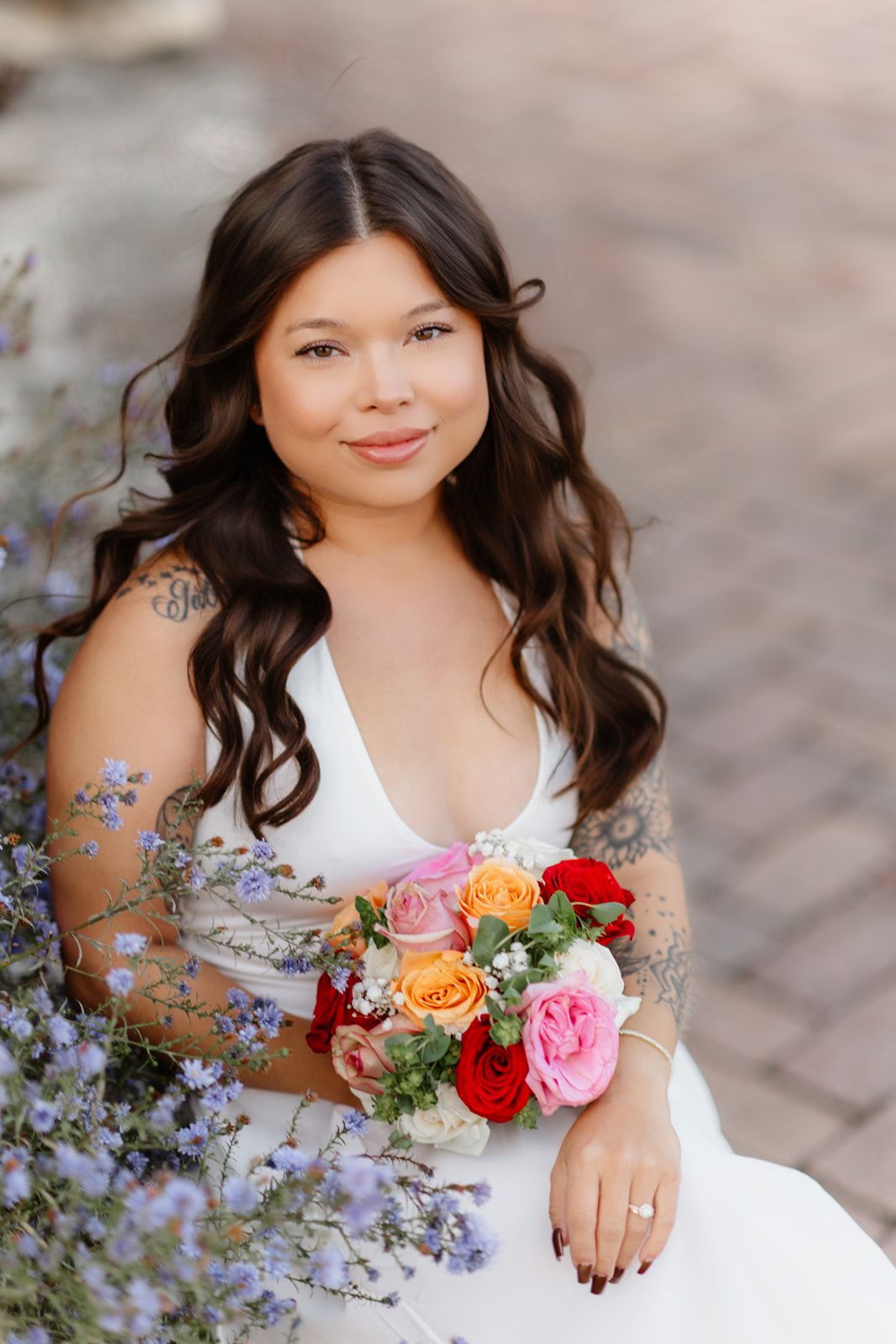 Bride in white gown holding colorful rose bouquet with tattoos visible, seated among purple wildflowers.