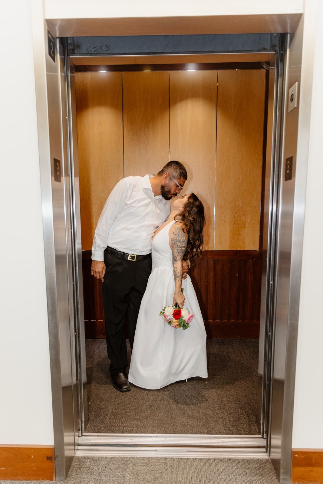 Newlyweds sharing a romantic kiss in a wood-paneled elevator, bride holding colorful rose bouquet.