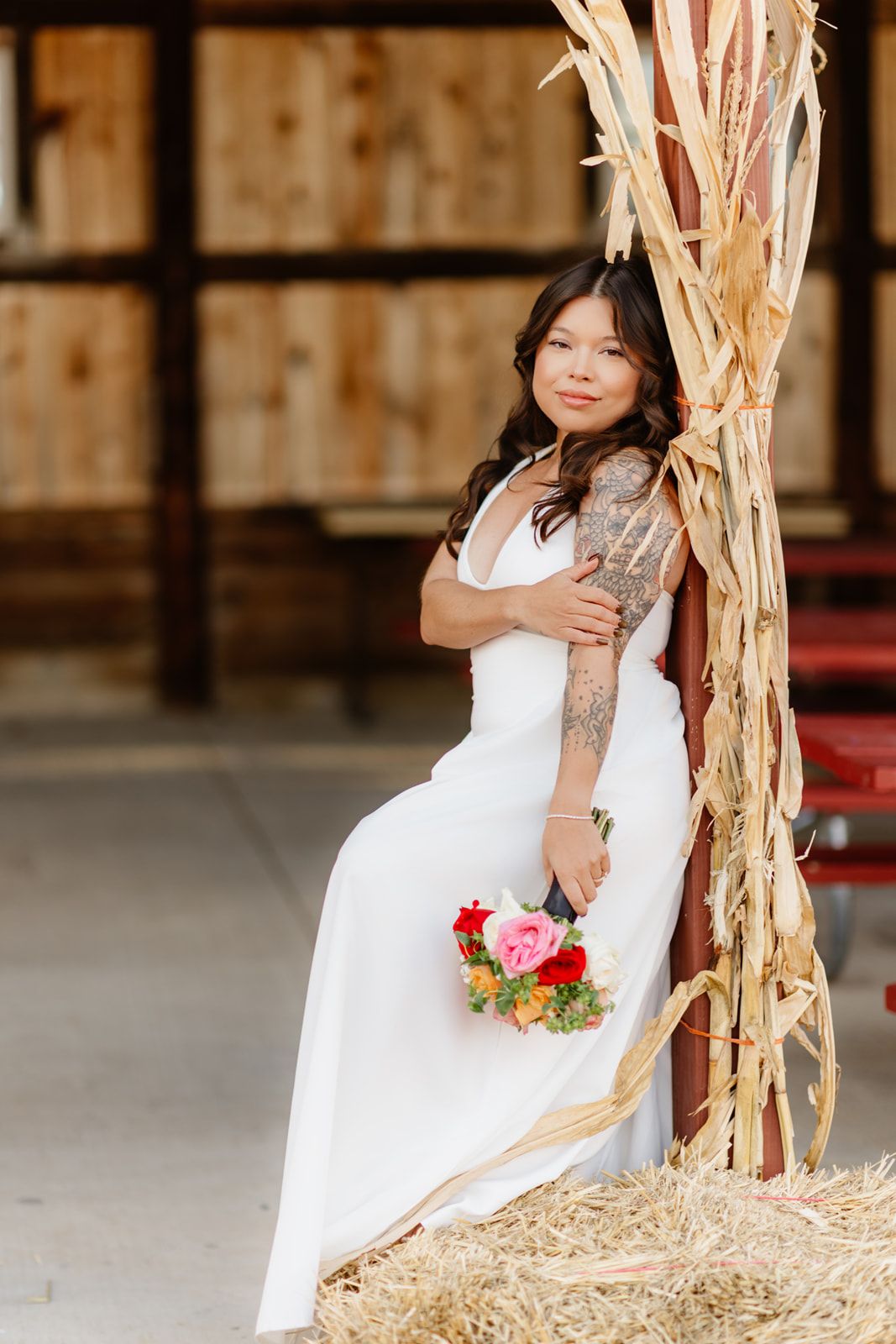Bride in white gown holding colorful bouquet poses by hay bales and dried corn stalks in a rustic barn setting.
