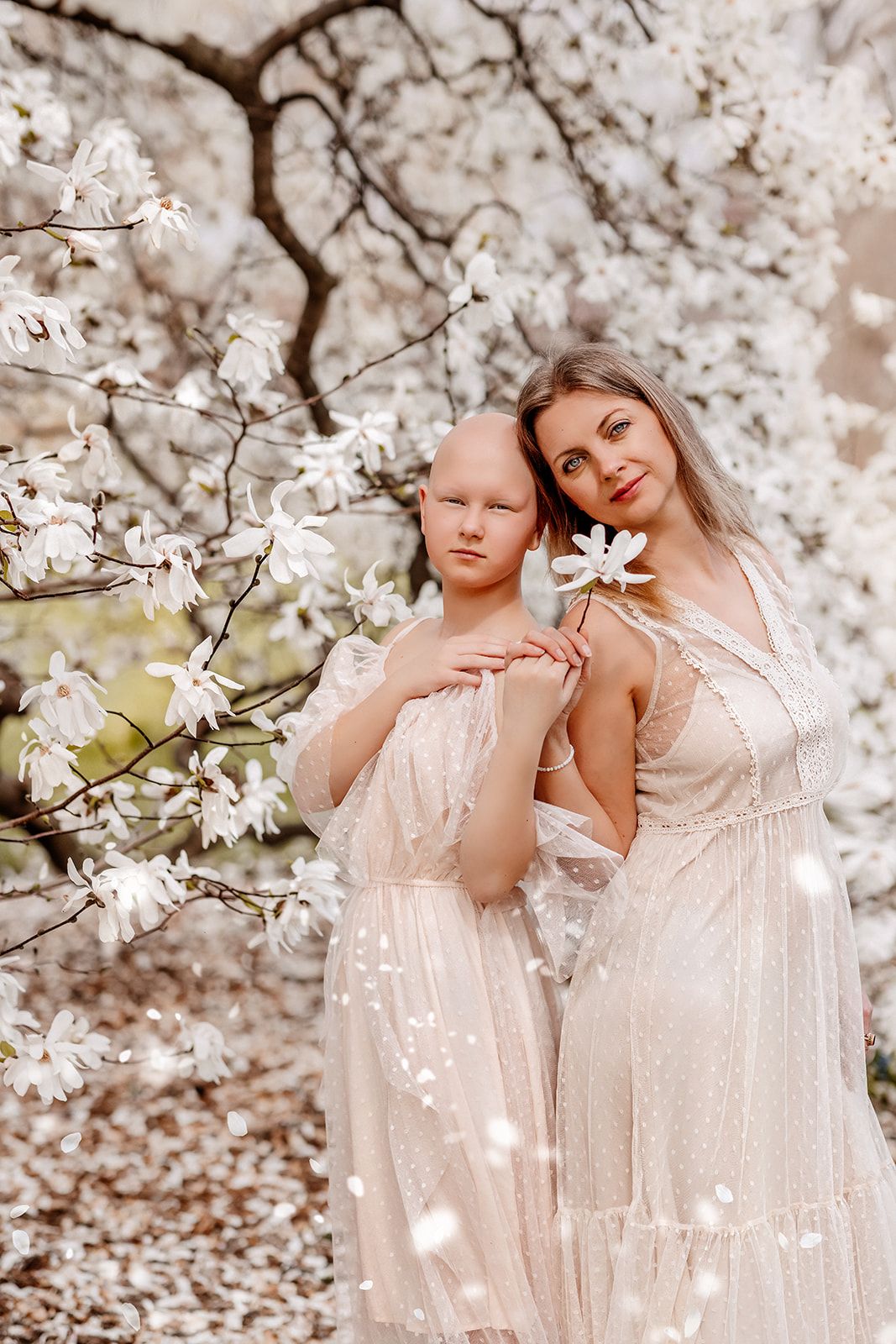 Two women in white dresses pose gracefully beneath blooming magnolia trees with white flowers.