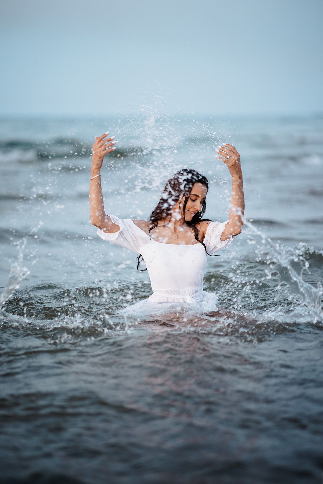 Woman in white dress splashing in ocean waves with arms raised.