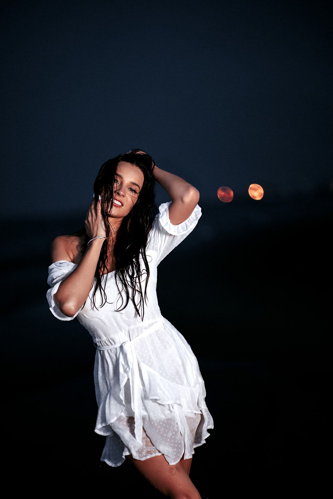 Woman in wet white dress poses on beach at night with bokeh lights in background.