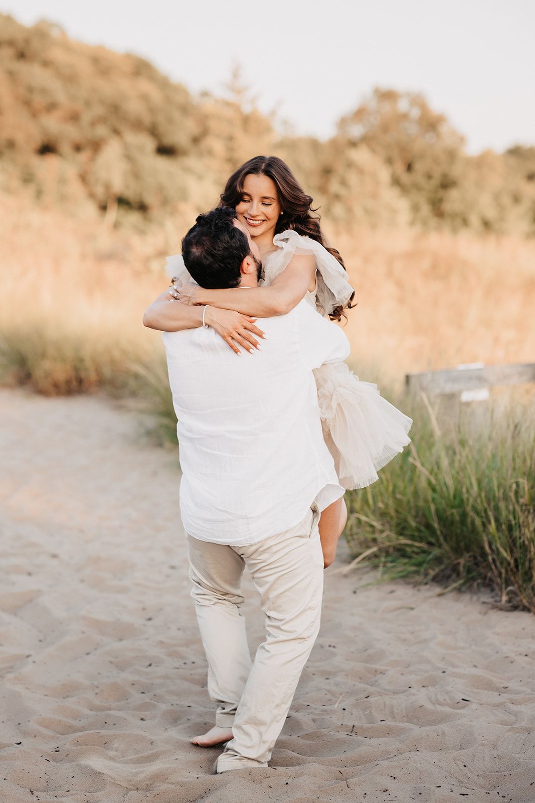 Man lifting smiling woman in white dress on sandy beach at warm golden sunset.