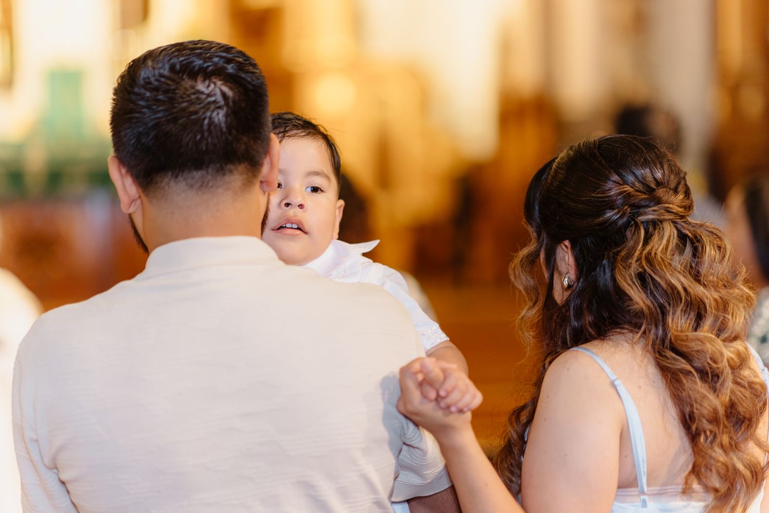 A father holds his baby in white christening outfit while mother looks on tenderly inside a church.