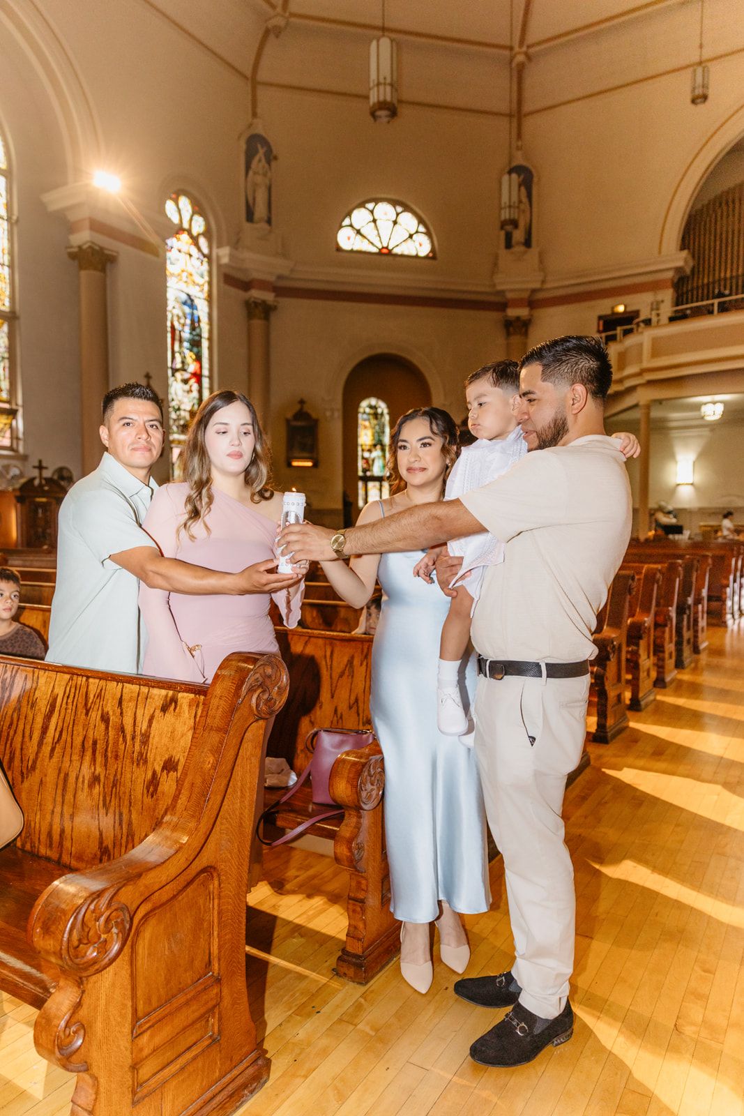 Family posing together inside an elegant church during a baptism, with stained glass windows and wooden pews in background.
