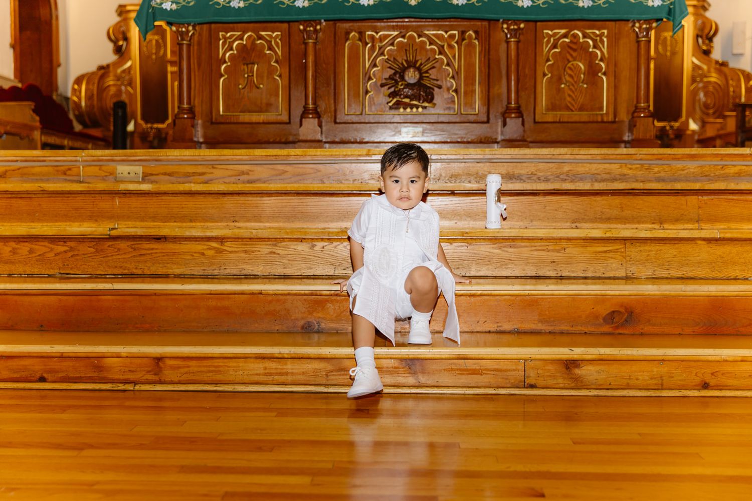 Toddler in white outfit sitting on wooden church steps holding a certificate during a baptism ceremony.