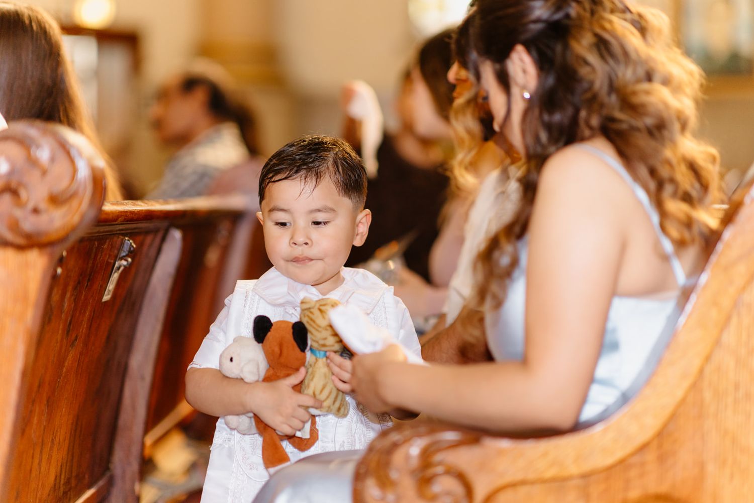 Young toddler in white baptism outfit holds stuffed animal toys while sitting in church pew.