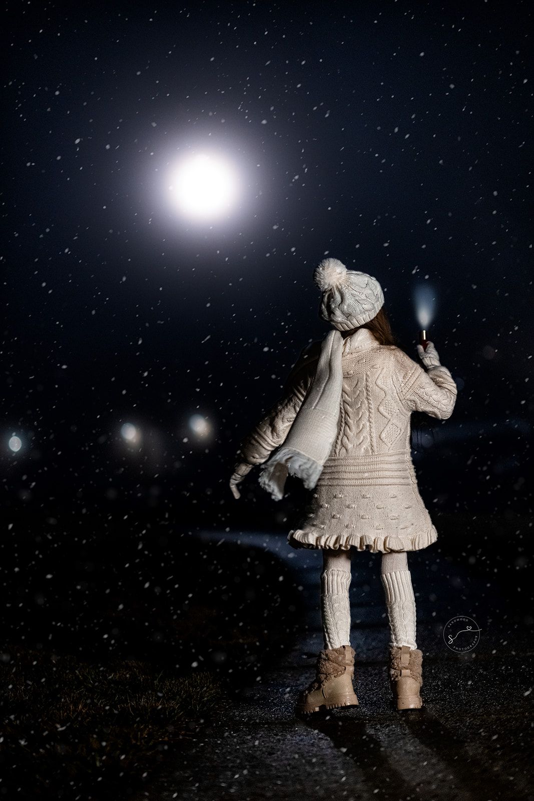 A girl in white winter knit outfit holds a flashlight under a glowing moon in a snowy night sky.