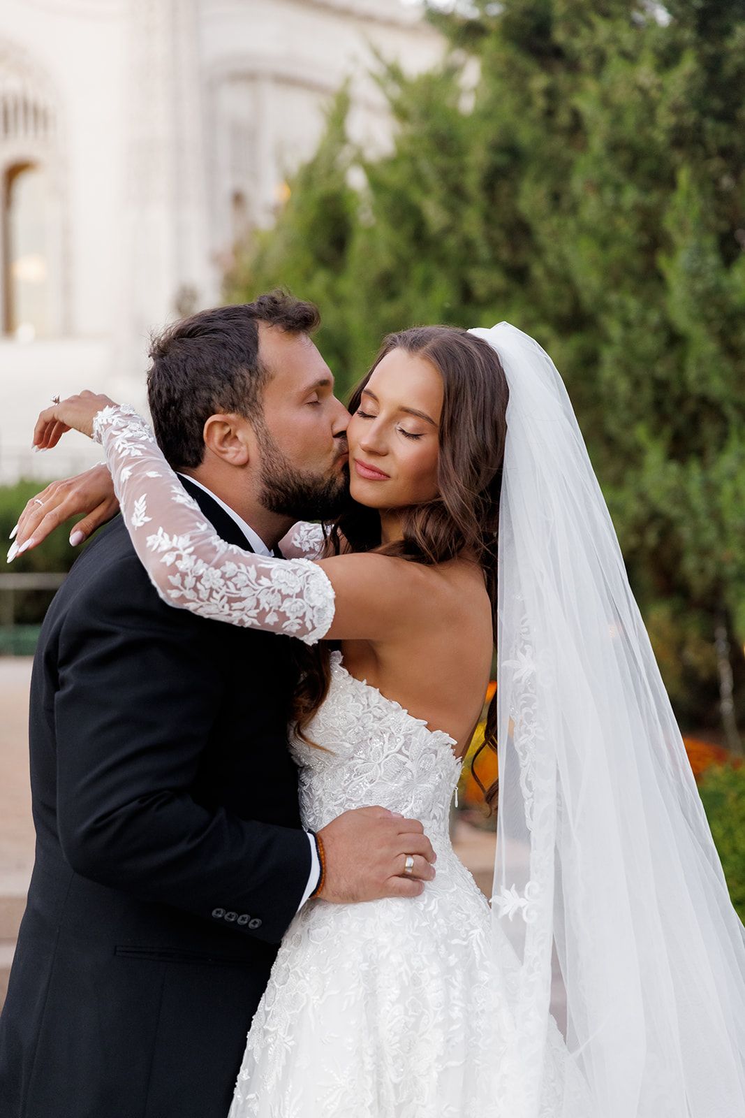 A groom in a black tuxedo kisses his bride in a lace wedding gown and veil outdoors near green trees.