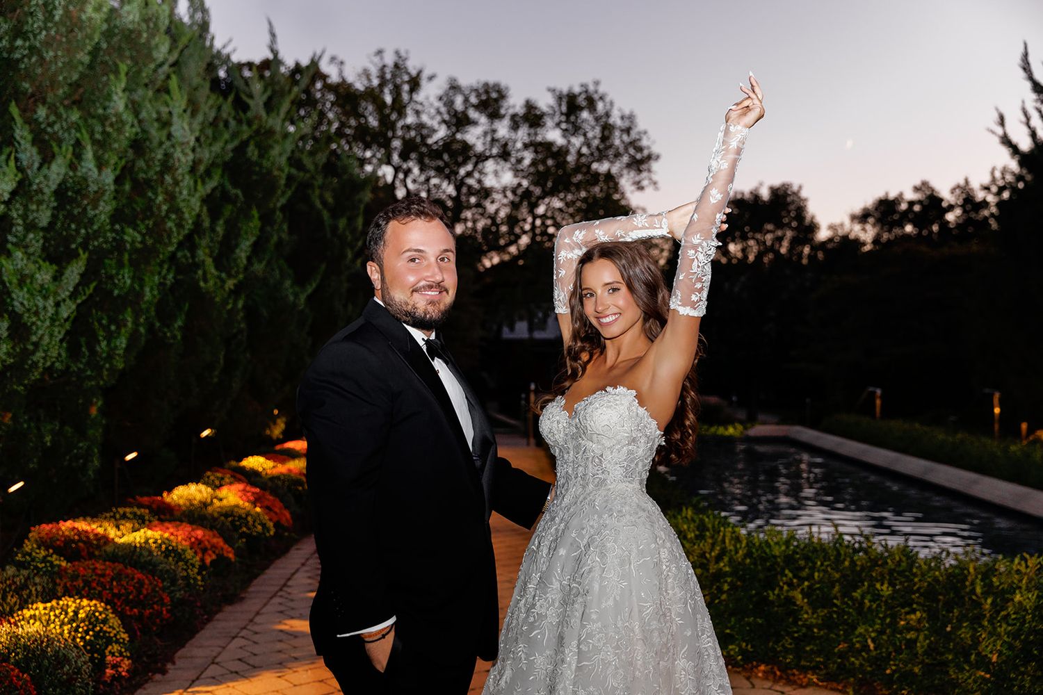 A couple in wedding attire poses outdoors at dusk, bride in sparkly gown with gloves, groom in black tuxedo.