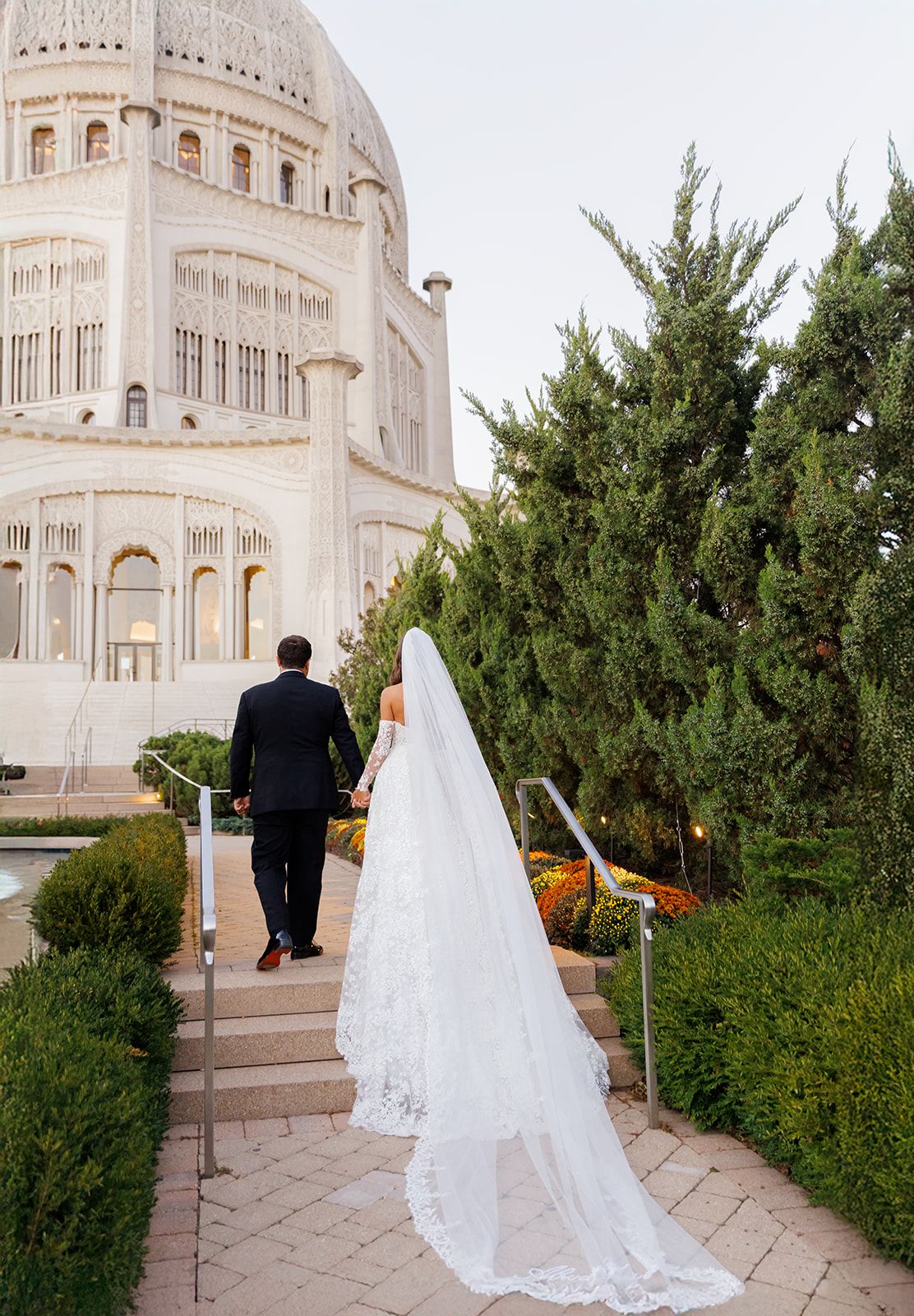 Bride in lace gown and cathedral veil walks with groom toward ornate white Bahai Temple in Wilmette, Illinois.