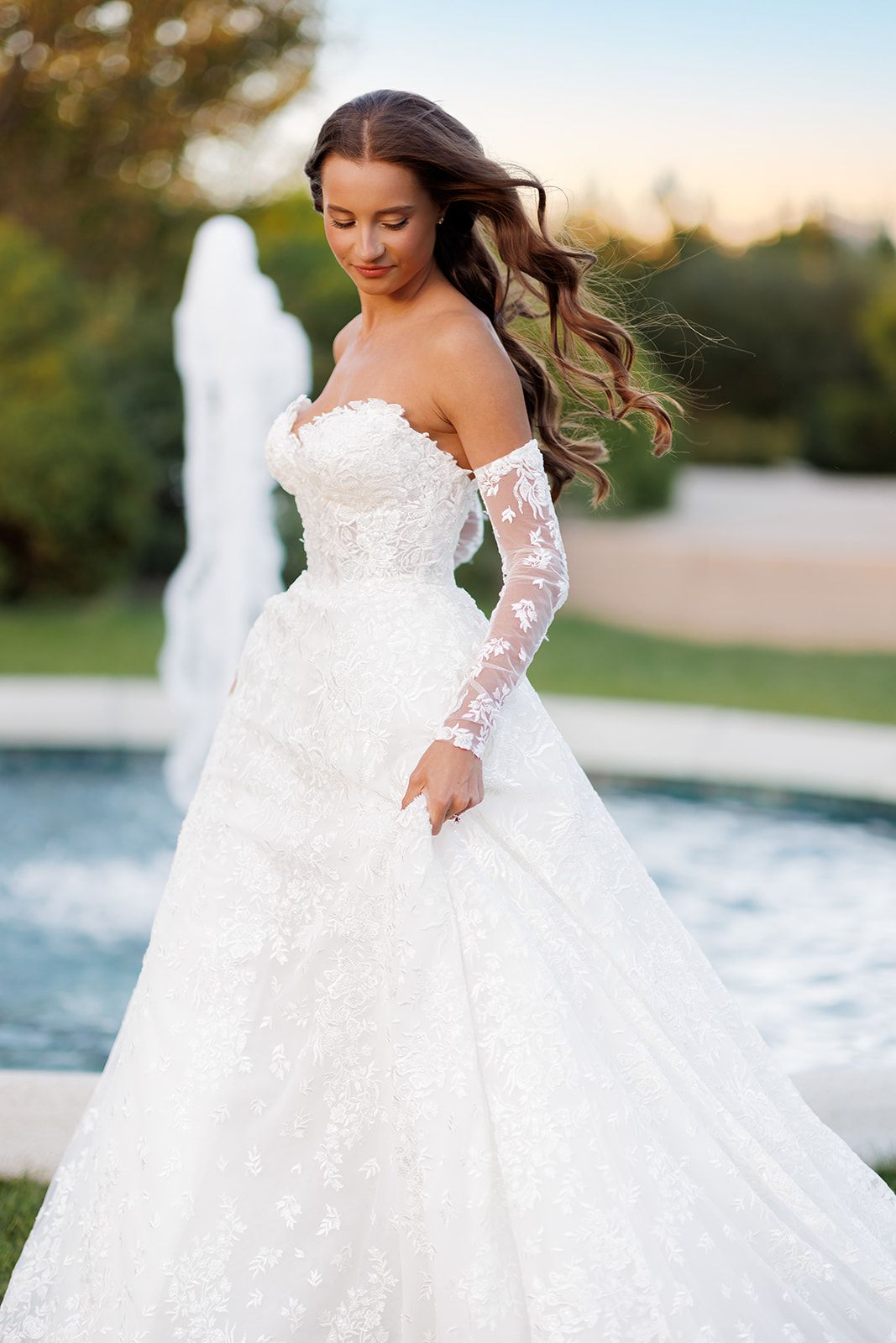 Bride in white lace off-shoulder ball gown with sheer sleeves, posing outdoors near a fountain.