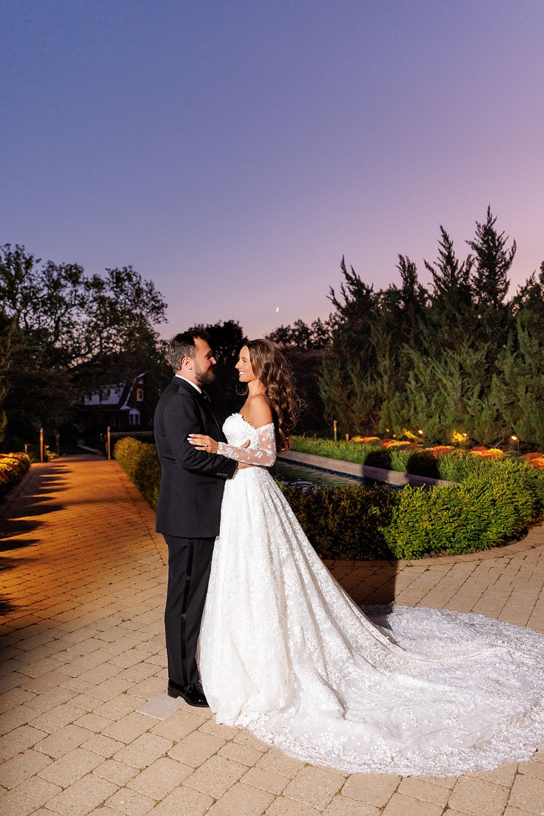 Bride and groom face each other tenderly on a lit garden path at sunset with cypress trees backdrop.