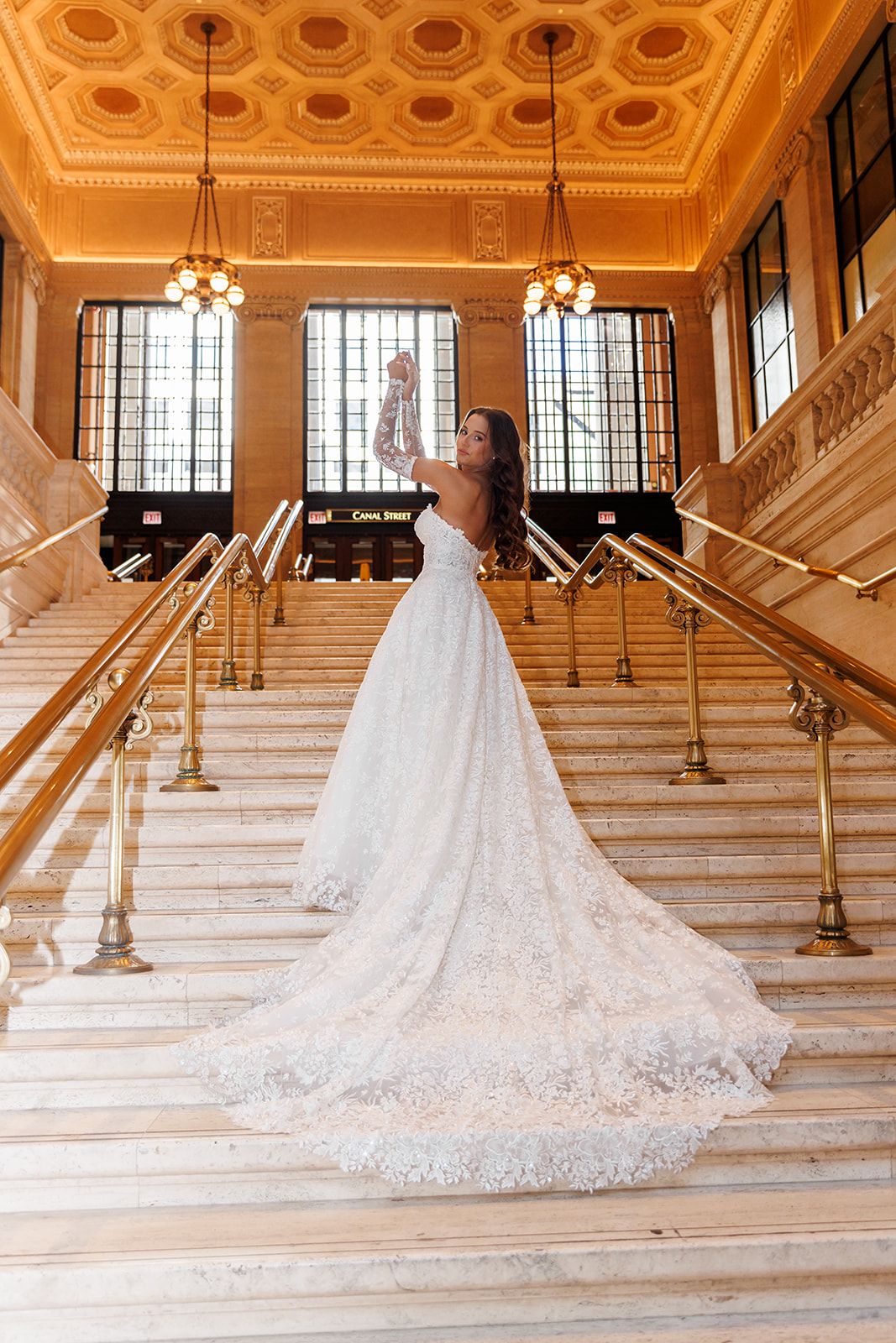 Bride in a stunning lace ball gown with a long train poses on marble stairs in an elegant golden hall.