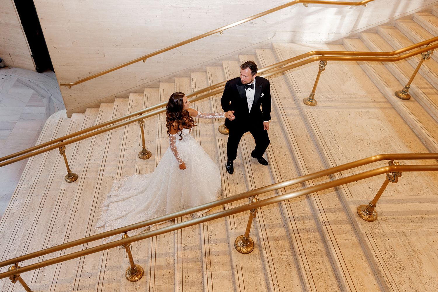 Aerial view of bride in white gown and groom in tuxedo ascending elegant staircase with gold railings.