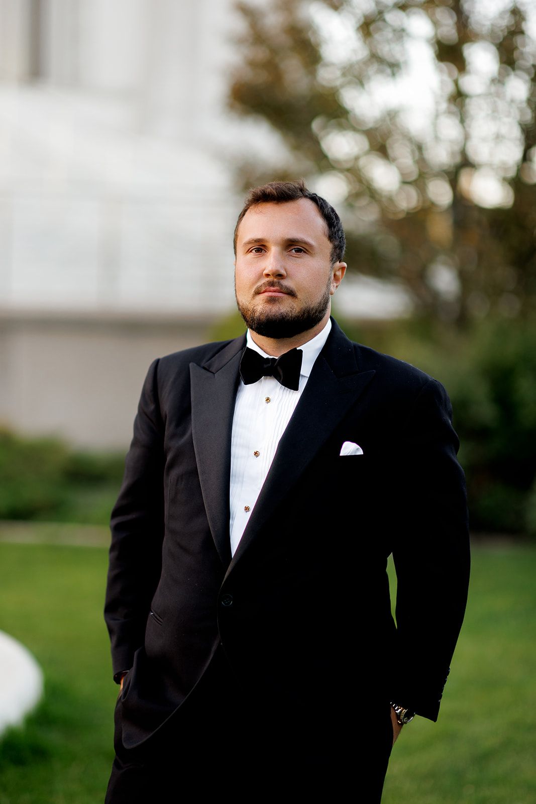 Elegant groom in black tuxedo with bow tie standing confidently outdoors on wedding day.