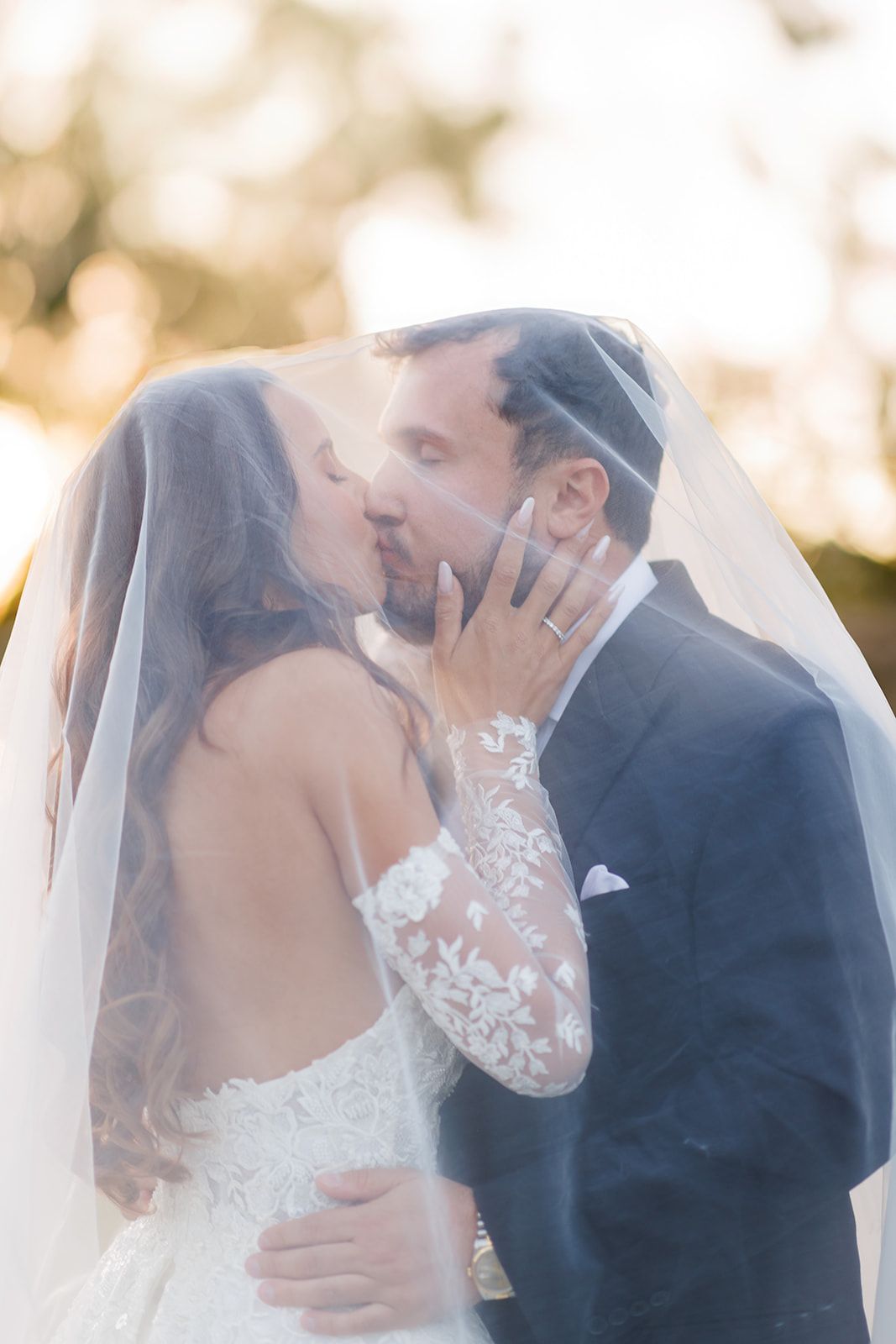 Bride and groom sharing a romantic kiss under a flowing veil, with soft golden bokeh background.