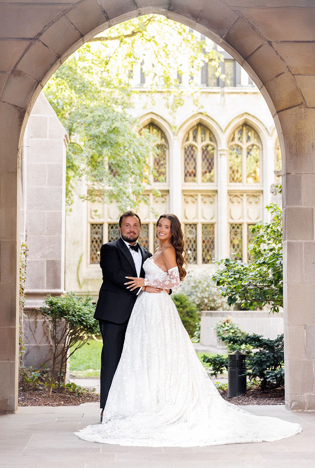 Bride and groom posing under a stone arch at a Gothic-style venue, bride in lace gown, groom in black tuxedo.