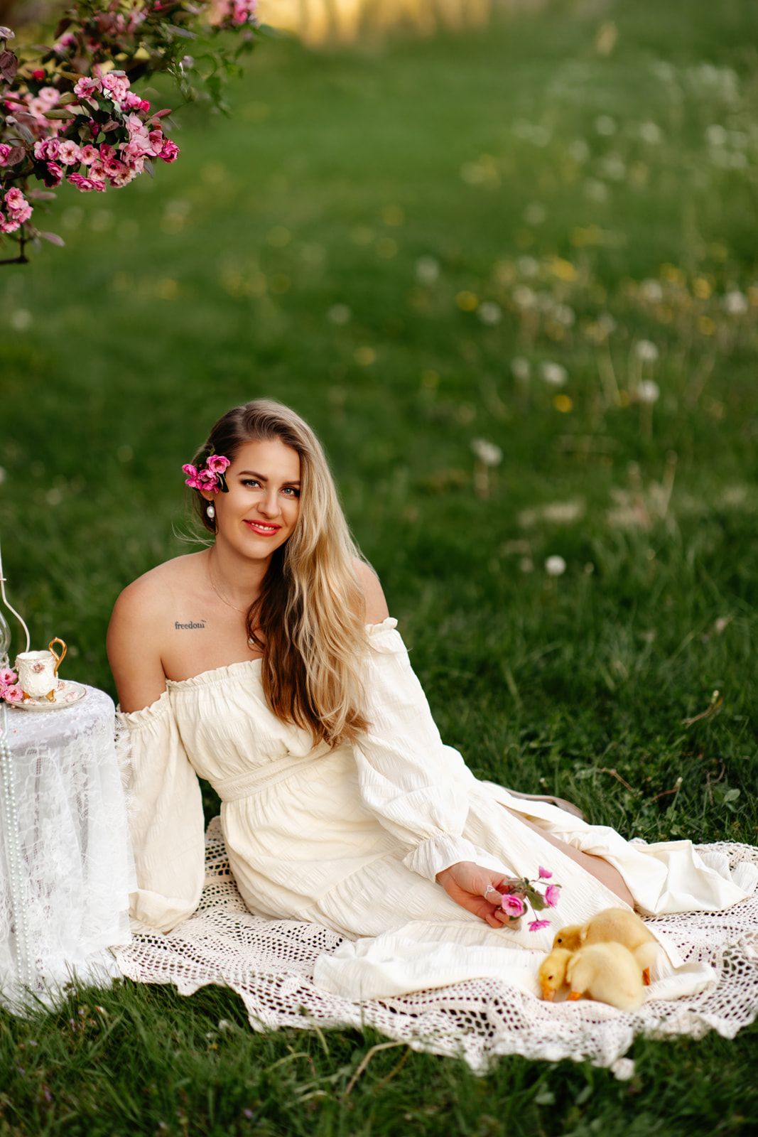 Blonde woman in white off-shoulder dress sits on lace blanket with ducklings near blooming pink flowers.
