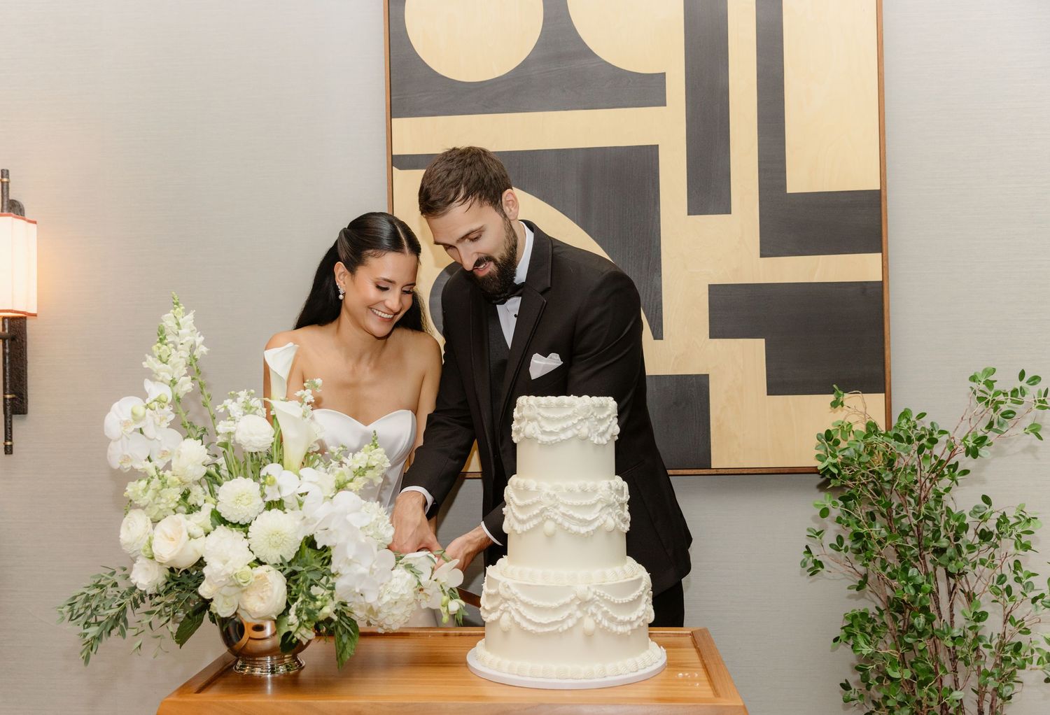 Smiling bride and groom cutting their white three-tiered ruffled wedding cake at elegant indoor reception.