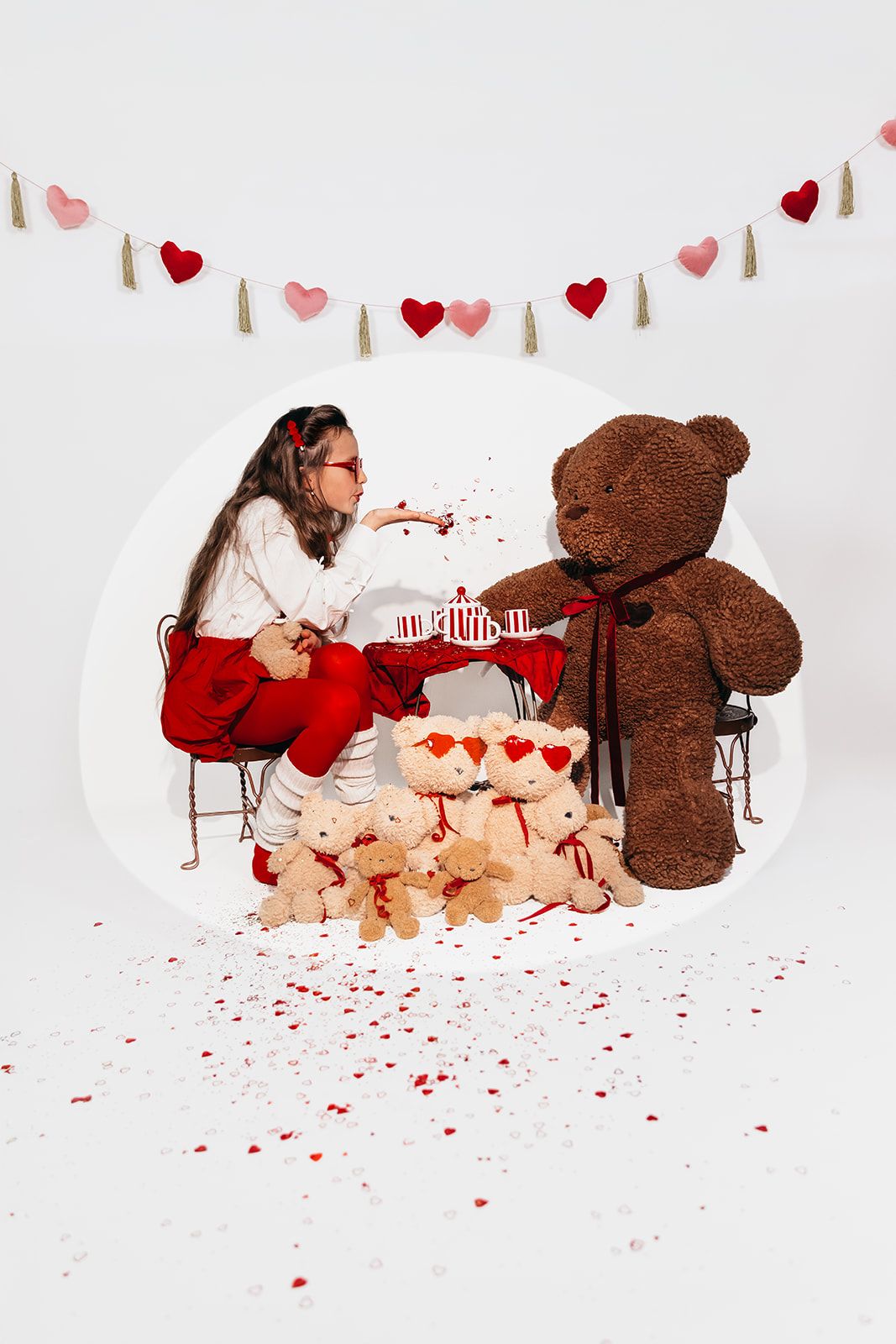 Girl in red pants has a tea party with a large brown teddy bear and small Valentine bears under heart garland.