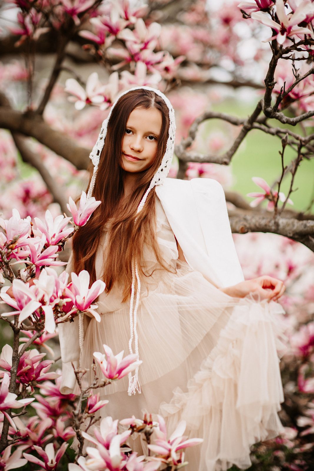Young girl in white dress and lace hood sits among pink magnolia blossoms in spring garden.