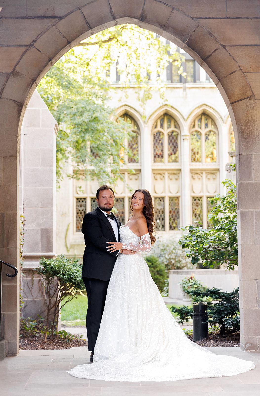Bride and groom pose under Gothic stone arch in wedding attire, lace gown with long train, black tuxedo.