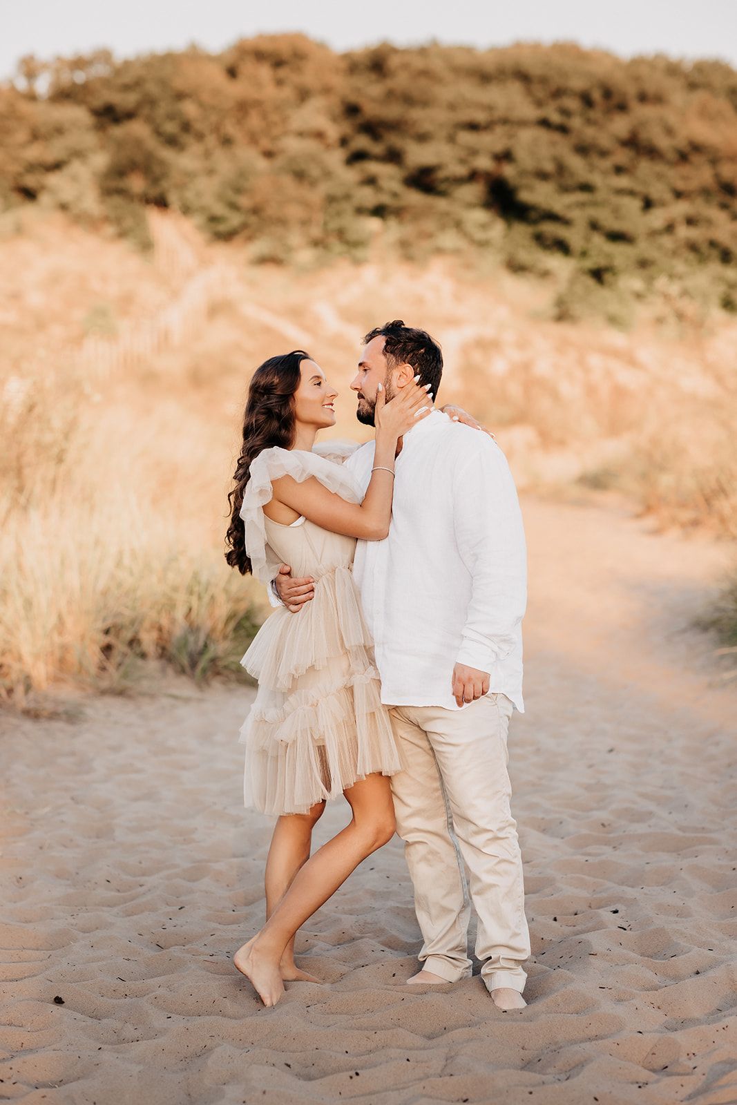 Couple in white outfits sharing a kiss on sandy beach dunes at golden hour.