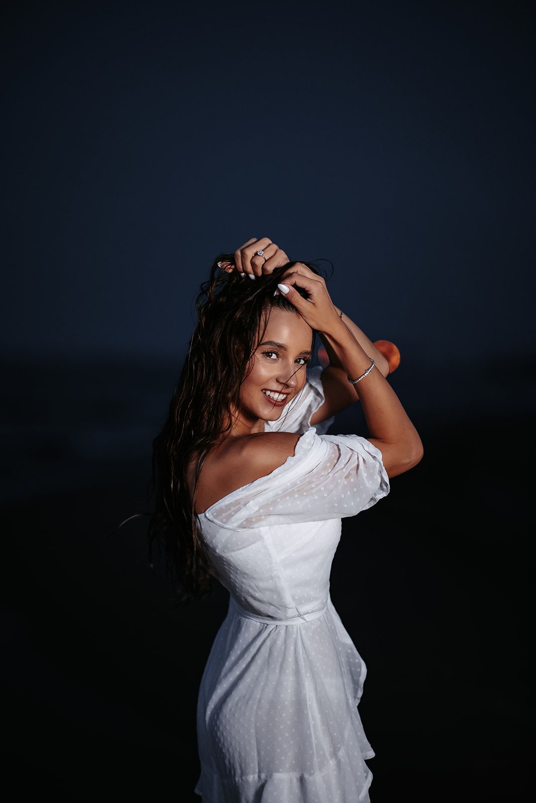 Smiling woman in white off-shoulder dress with wet hair at night.