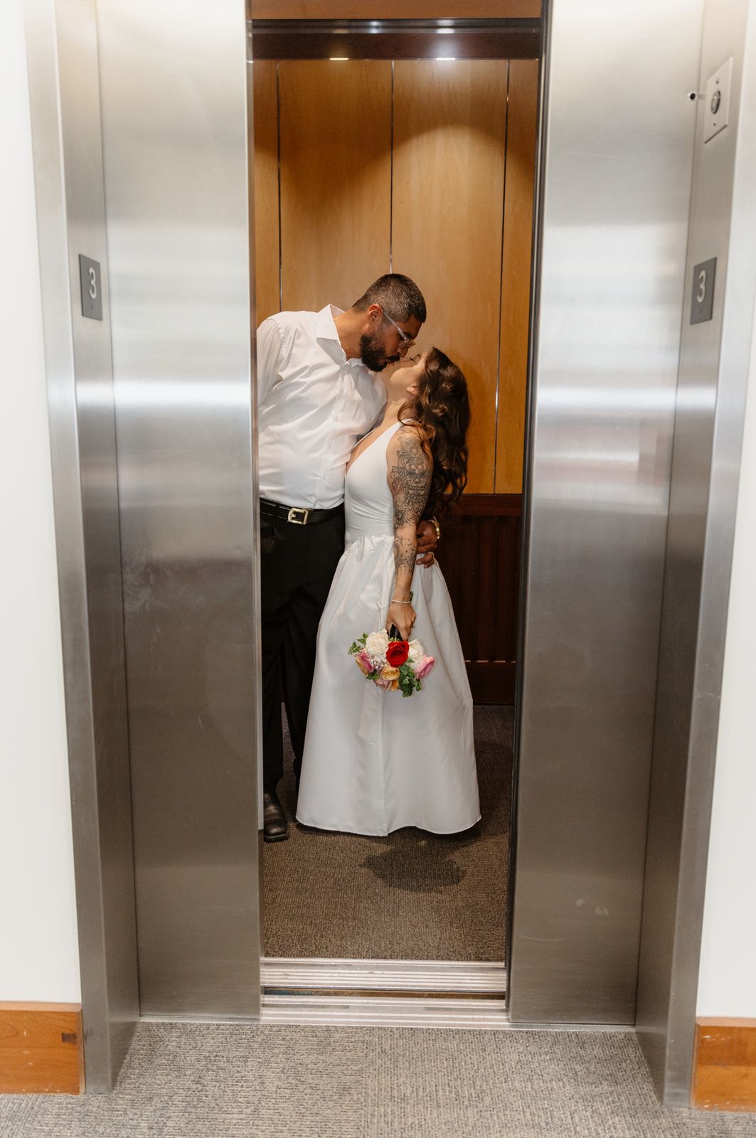 Newlywed couple sharing a romantic kiss in an elevator, bride in white gown holding colorful bouquet, groom in white shirt.