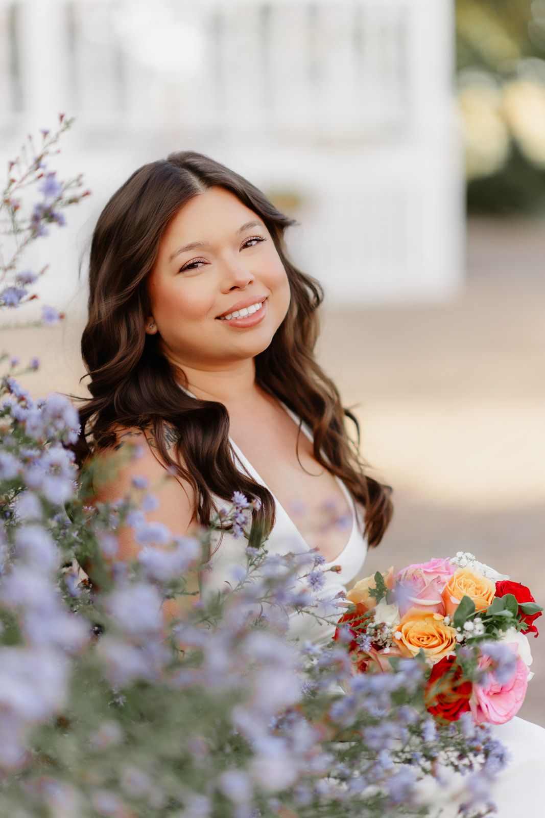 Smiling bride with dark wavy hair holds colorful bouquet of roses surrounded by purple wildflowers outdoors.