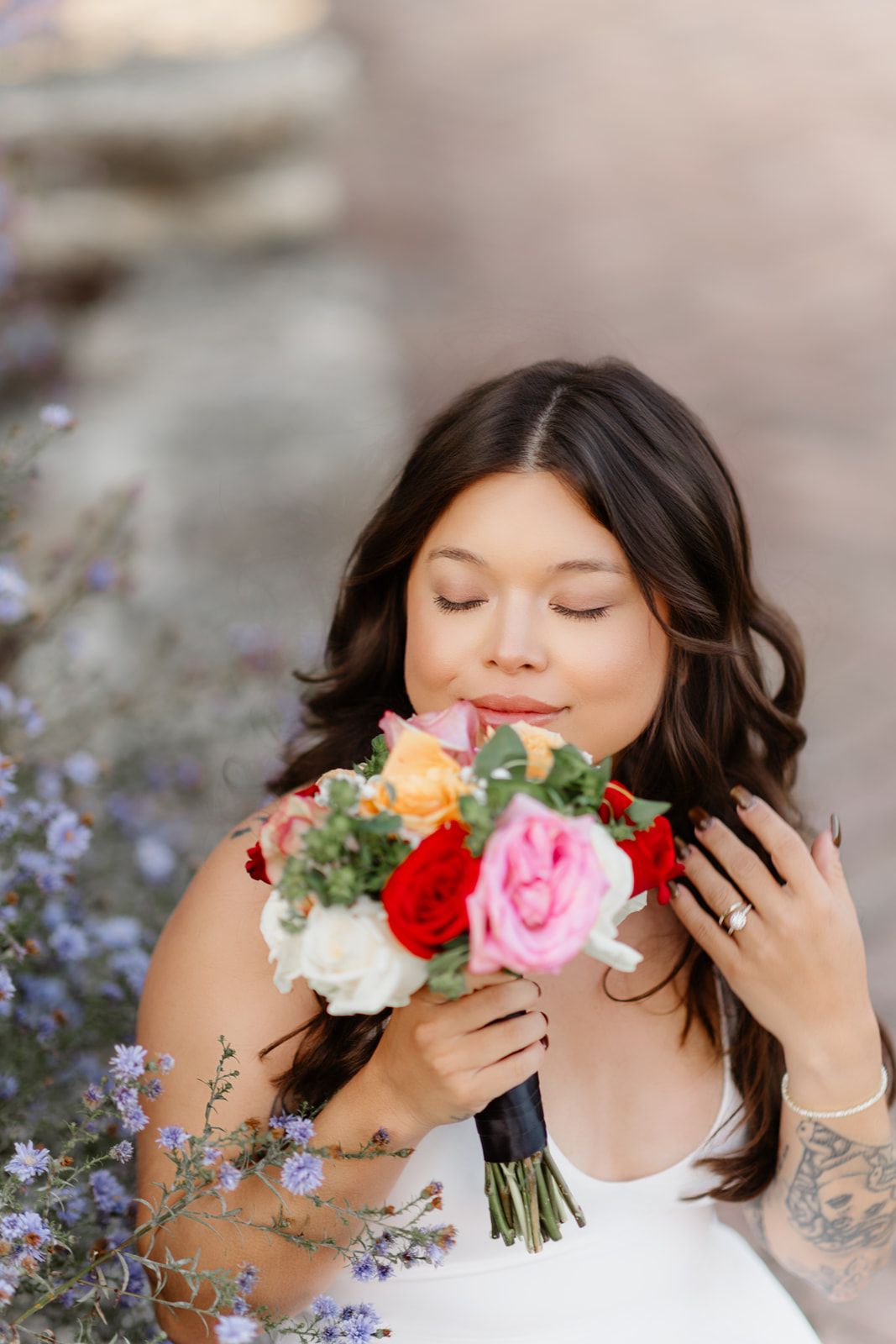 Bride with dark hair smells colorful rose bouquet outdoors, eyes closed, wearing white dress and ring.