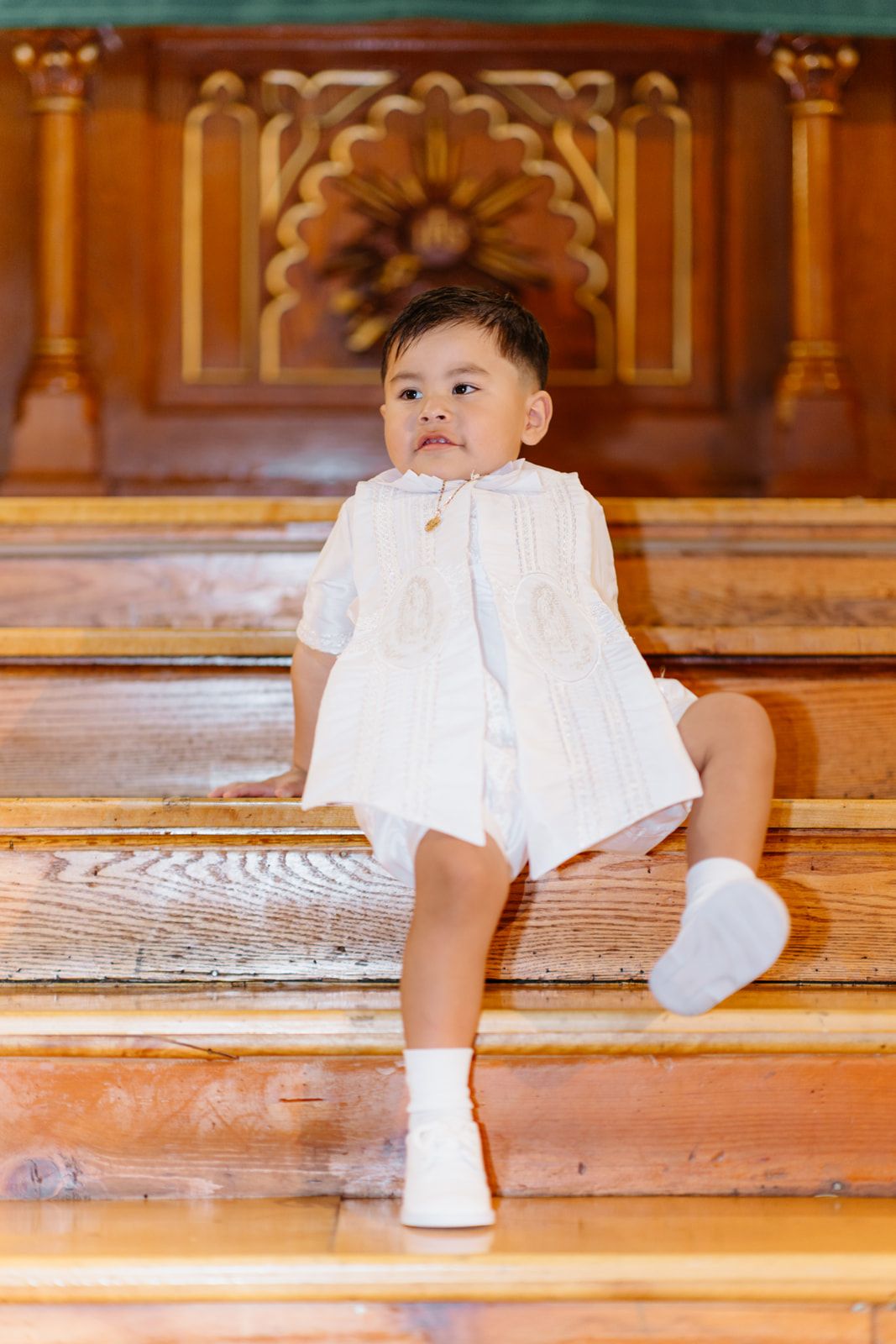 Adorable baby in white outfit sitting on wooden church steps, looking sideways with curiosity.