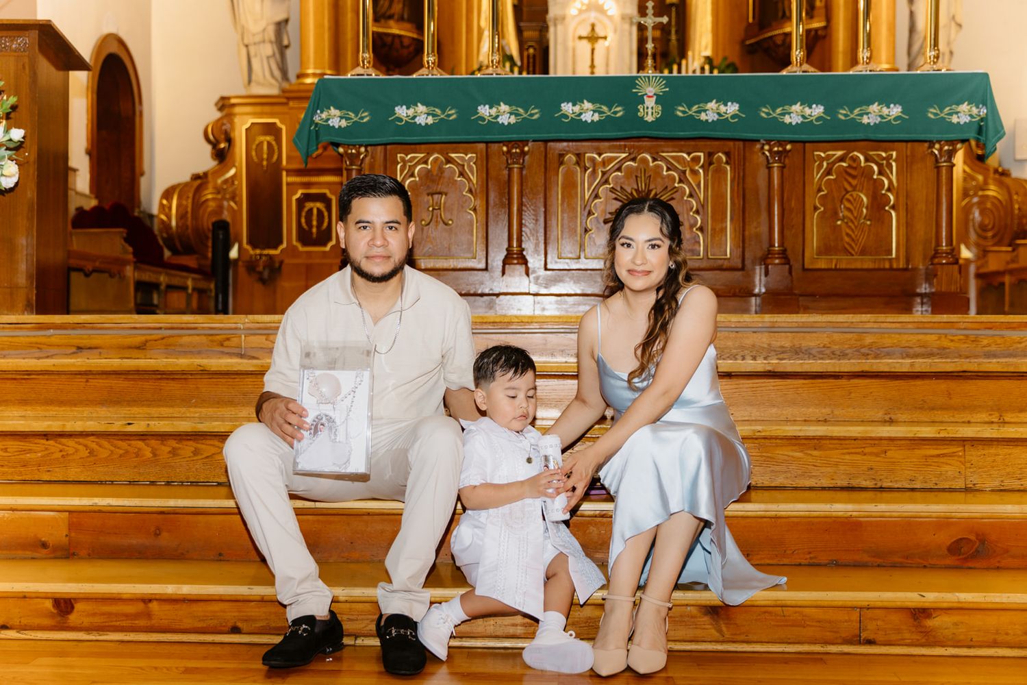 Family of three in white and blue outfits posing on church steps during a baptism celebration.