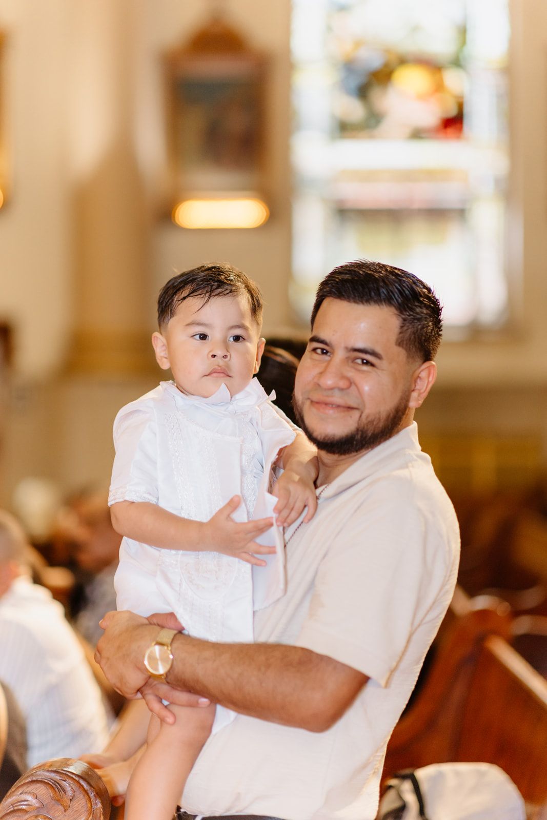 Man with beard holds toddler boy in white outfit inside a church during a baptism ceremony.