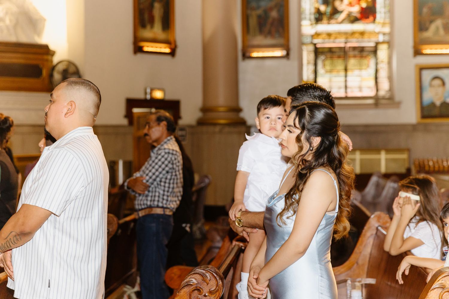 A mother holds her young son in a sunlit church during a baptism ceremony, surrounded by family in wooden pews.