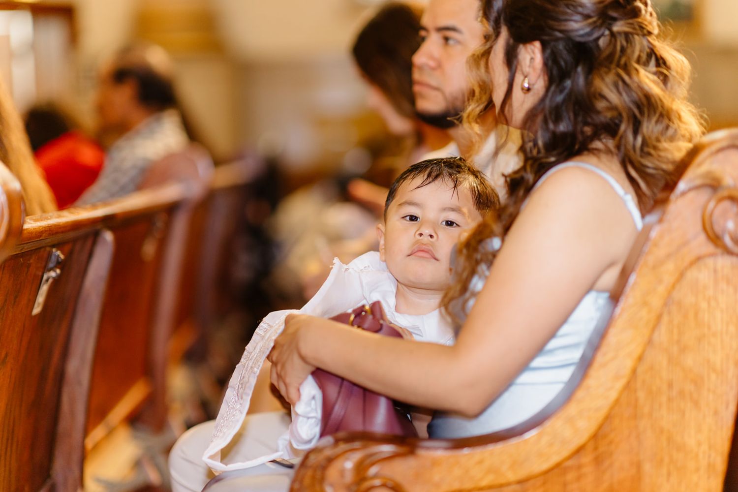 A young child in white sits on a woman's lap in a church pew during a wedding ceremony.
