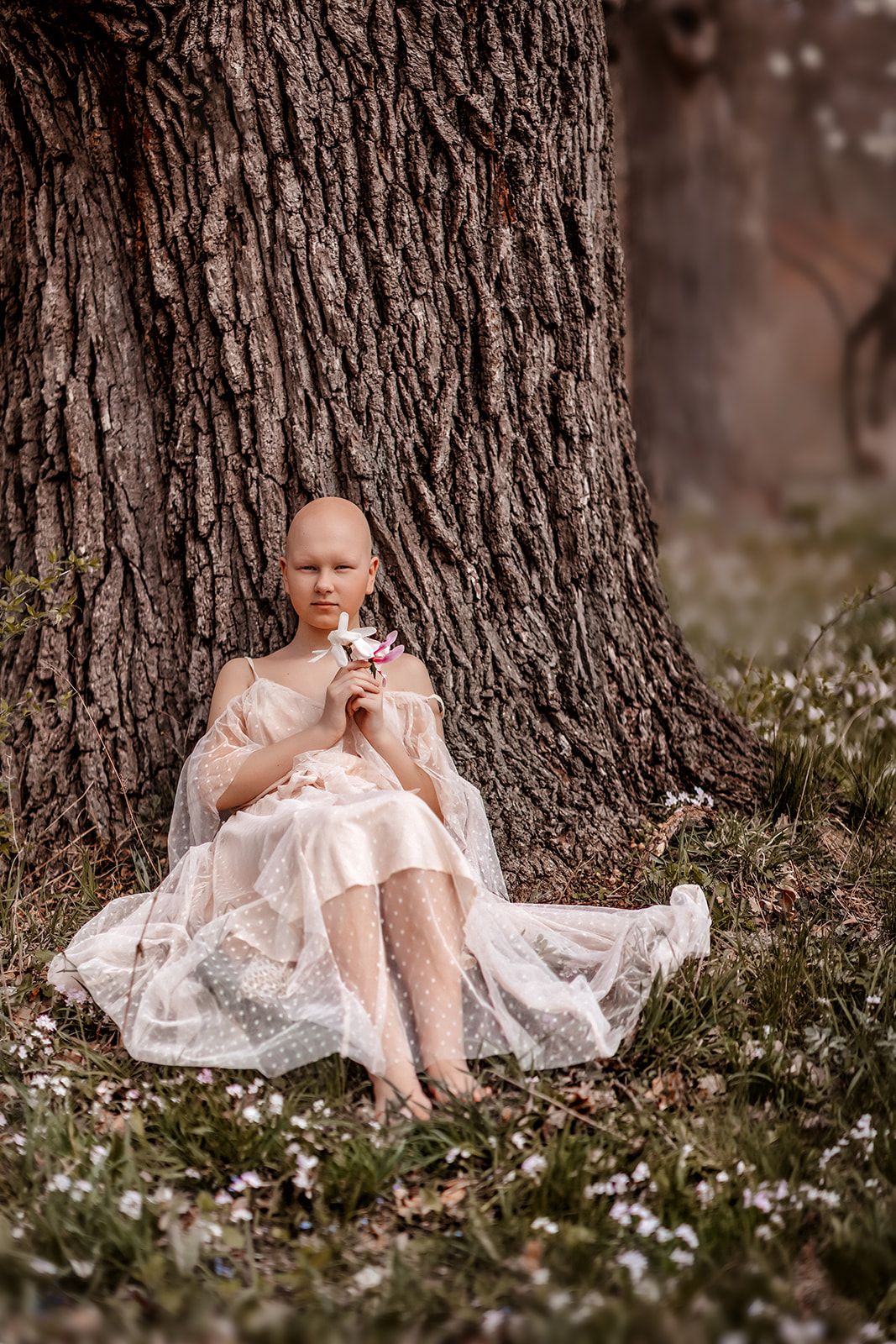 Bald woman in white lace dress sitting against tree trunk holding flower in blooming garden.