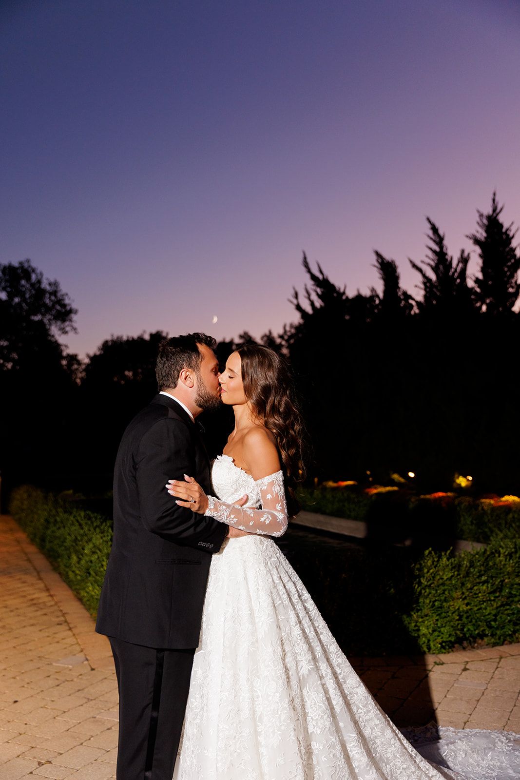 Couple shares an intimate kiss on a garden pathway under a purple twilight sky with tall trees.