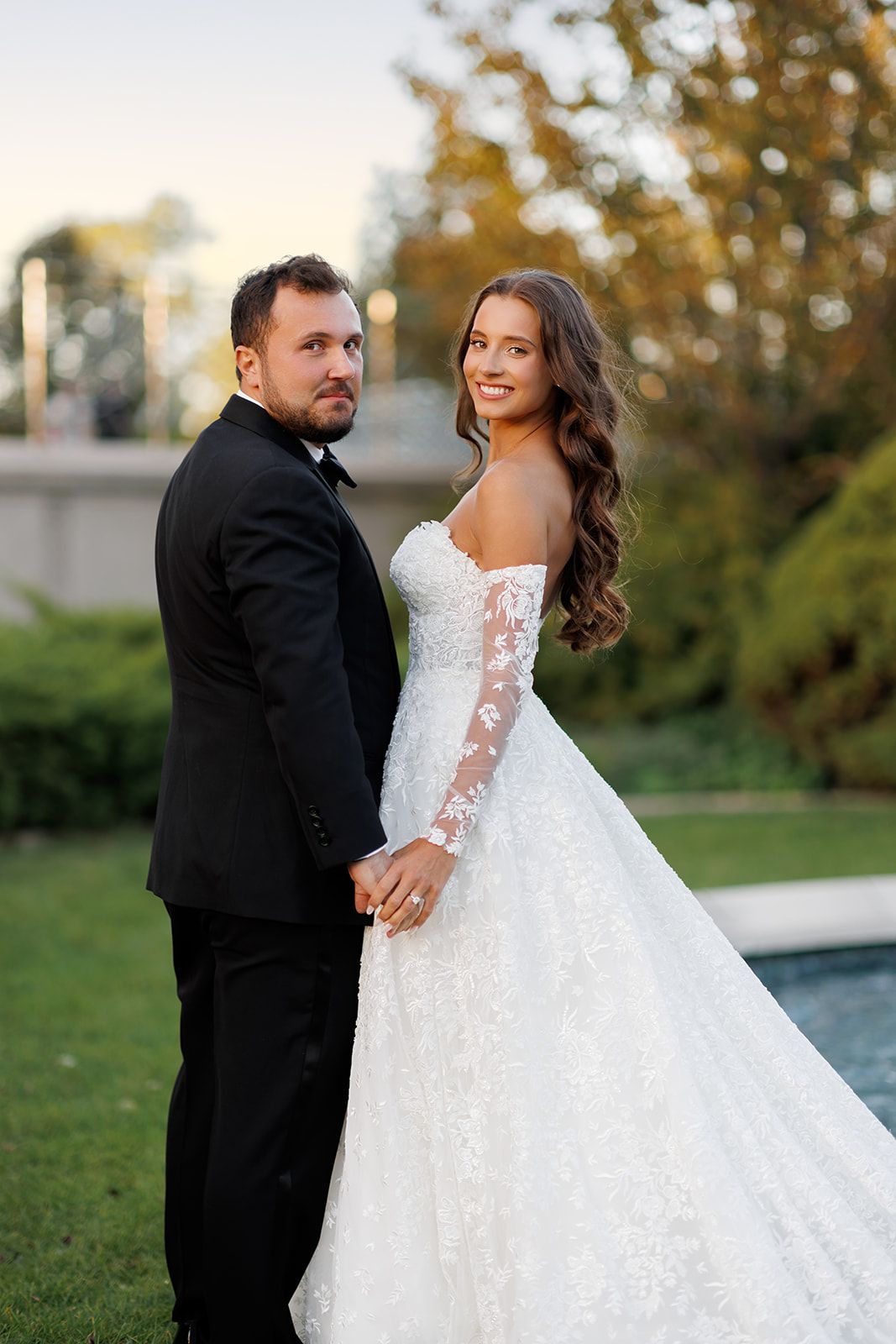 A bride in a lace off-shoulder gown holds hands with a groom in a black tuxedo outdoors near a pool.