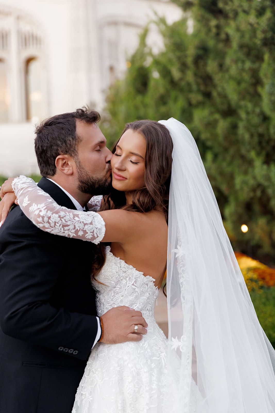 Newlyweds embrace as groom kisses bride on the cheek, bride in lace wedding dress with flowing veil in garden setting.
