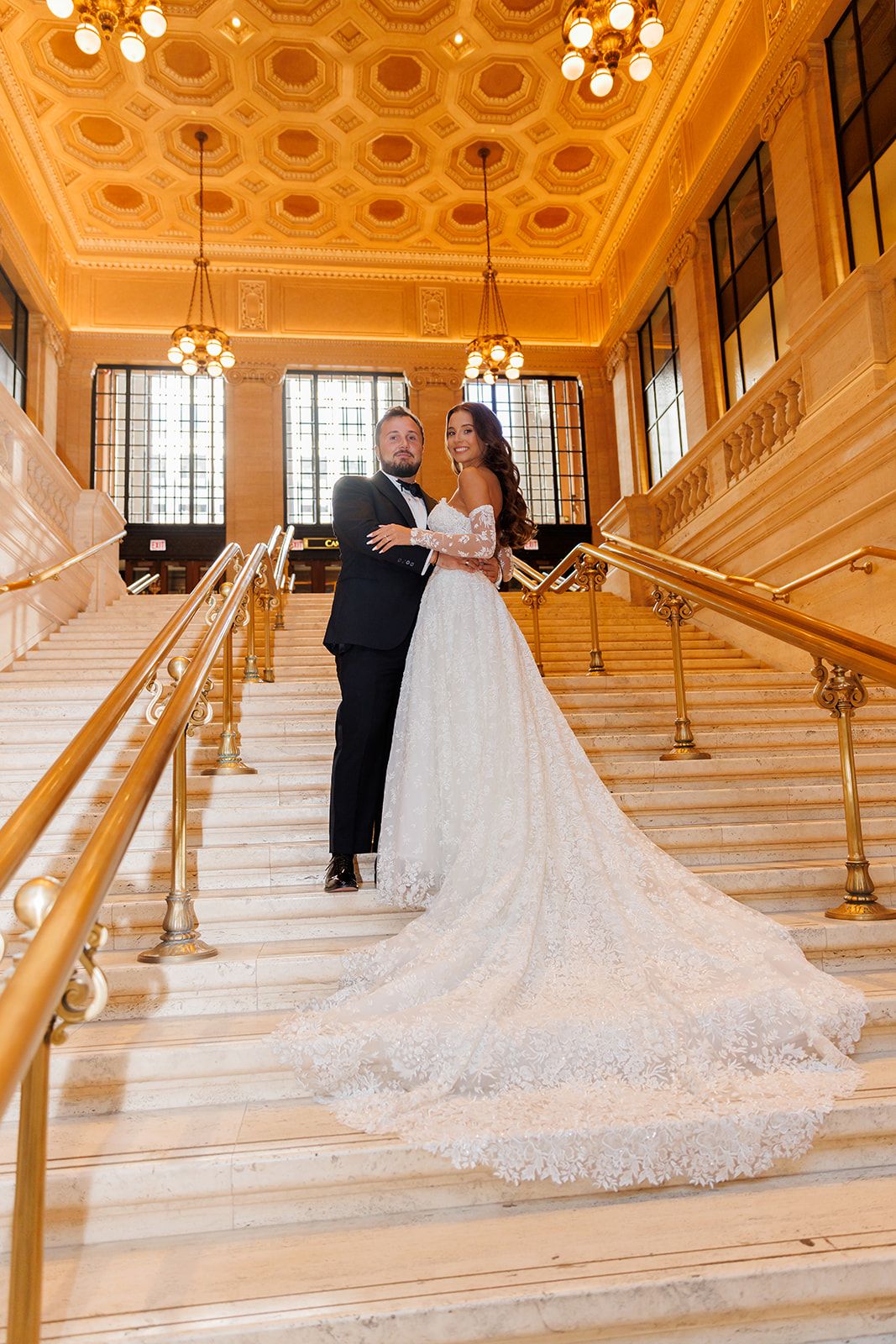 Bride and groom pose on grand marble staircase with gold railings in an elegant ballroom venue.