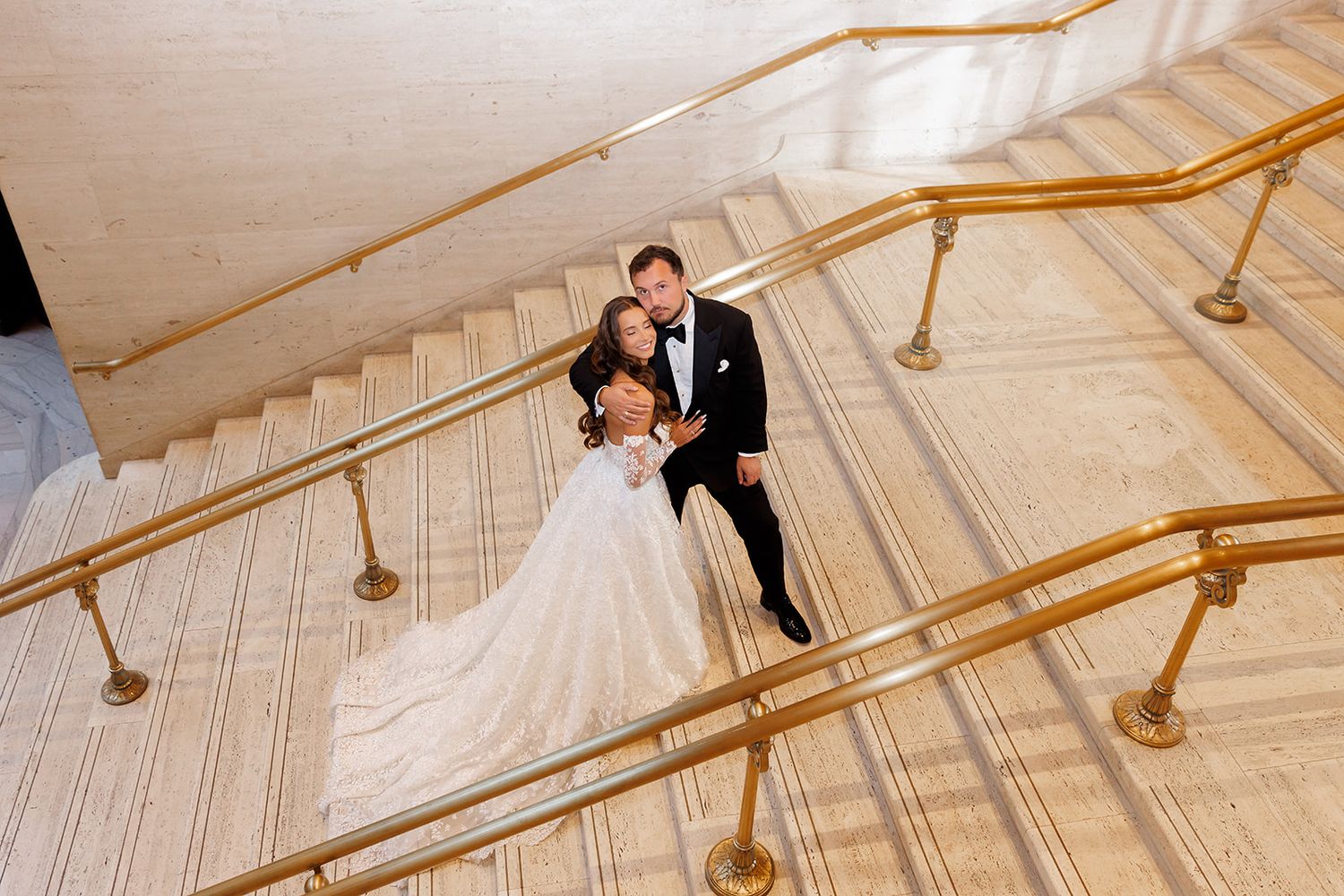Bride and groom share a romantic moment on a grand marble staircase with gold railings at an elegant wedding venue.