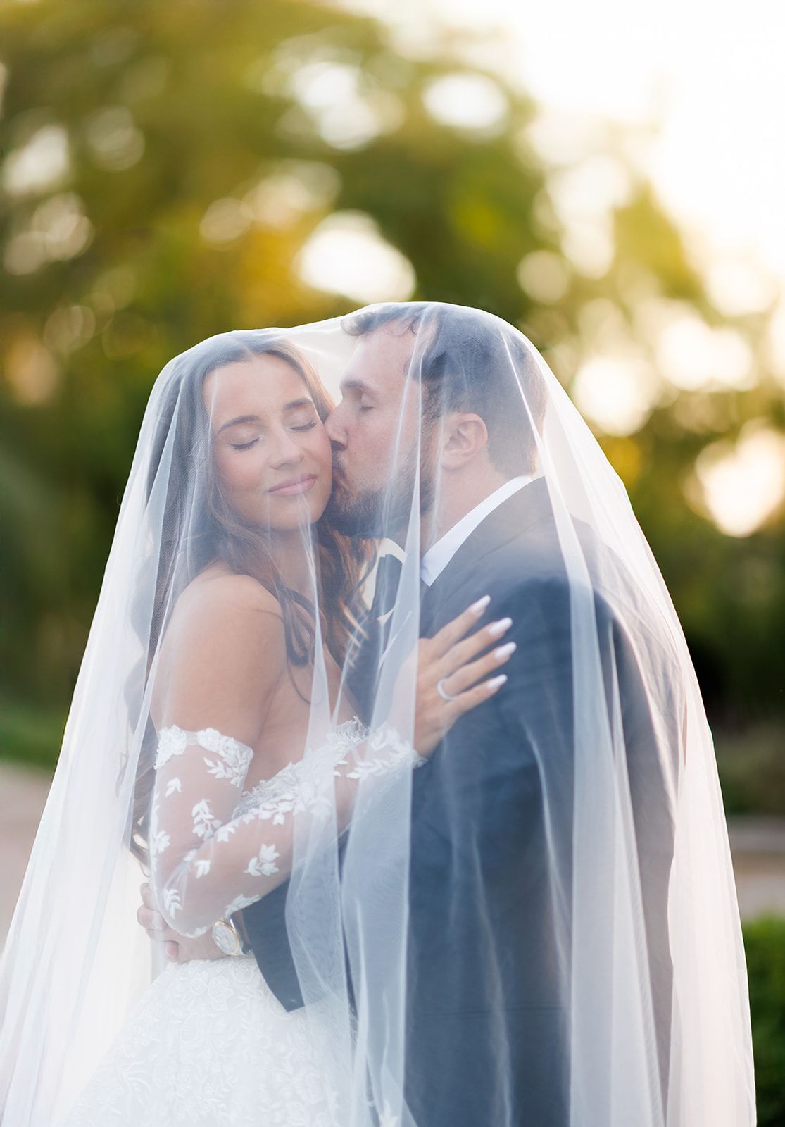 Bride and groom sharing a romantic kiss under a flowing white veil at golden hour during their outdoor wedding.