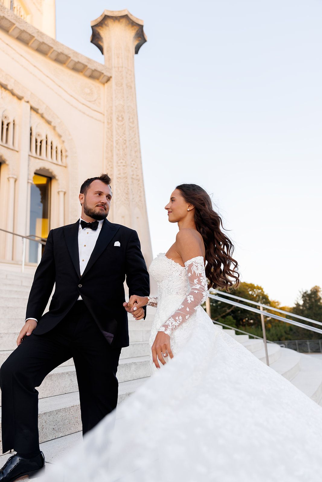 Bride and groom hold hands walking up grand stone steps of a beautiful wedding venue.